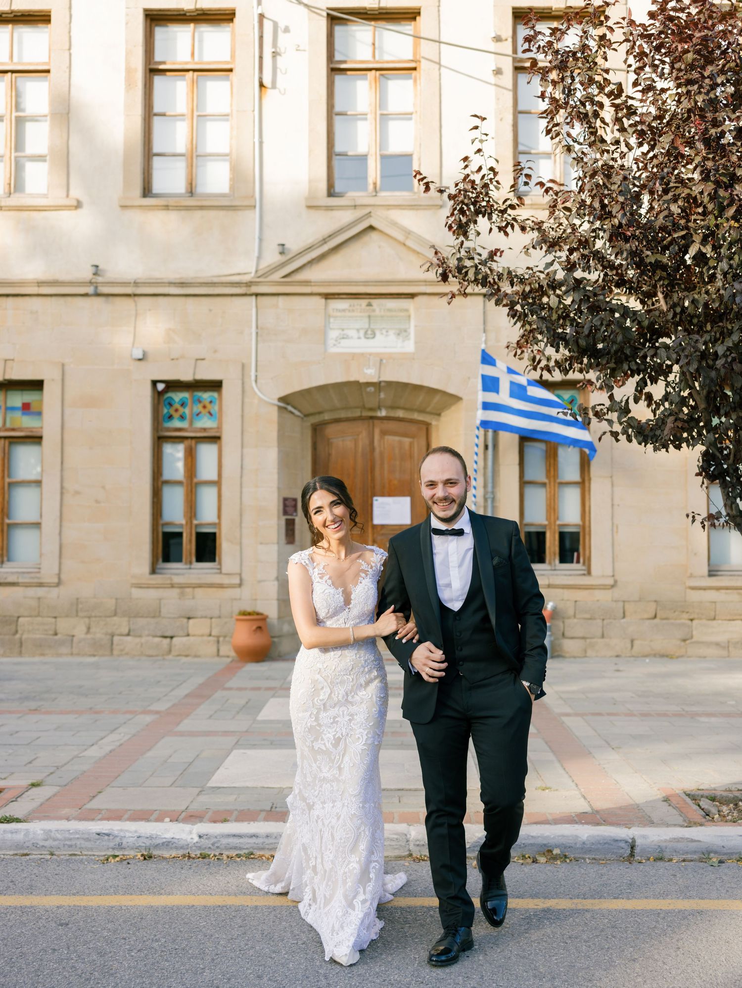Newlyweds strolling and laughing outside historic stone building with Greek flag.