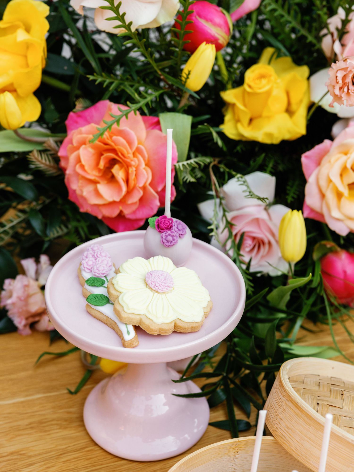 Pink cake stand displays delicate butterfly sugar cookies with floral arrangements of yellow and pink roses in background.