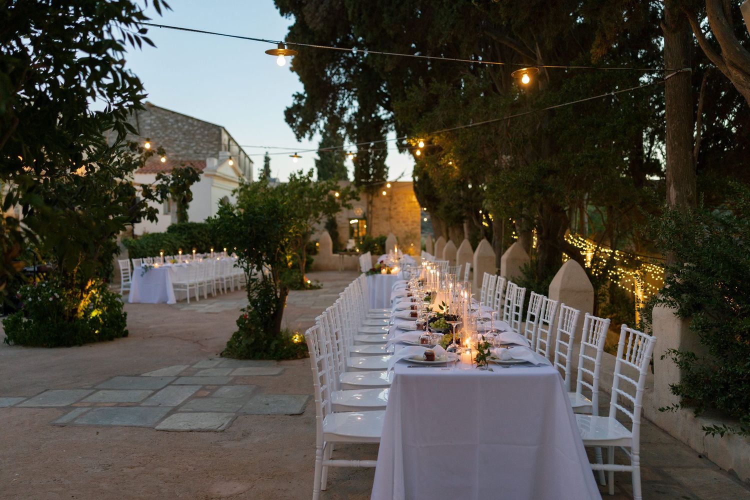 Elegant outdoor wedding reception with long white-draped tables, chiavari chairs, and string lights at dusk.