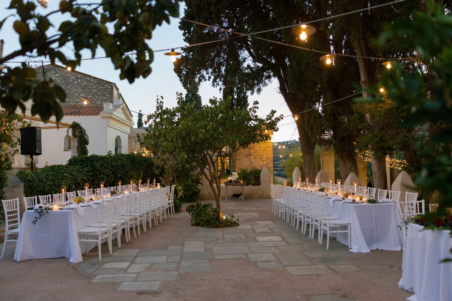Elegant outdoor wedding reception with long white-draped tables, string lights, and lush trees at a rustic stone venue at dusk.