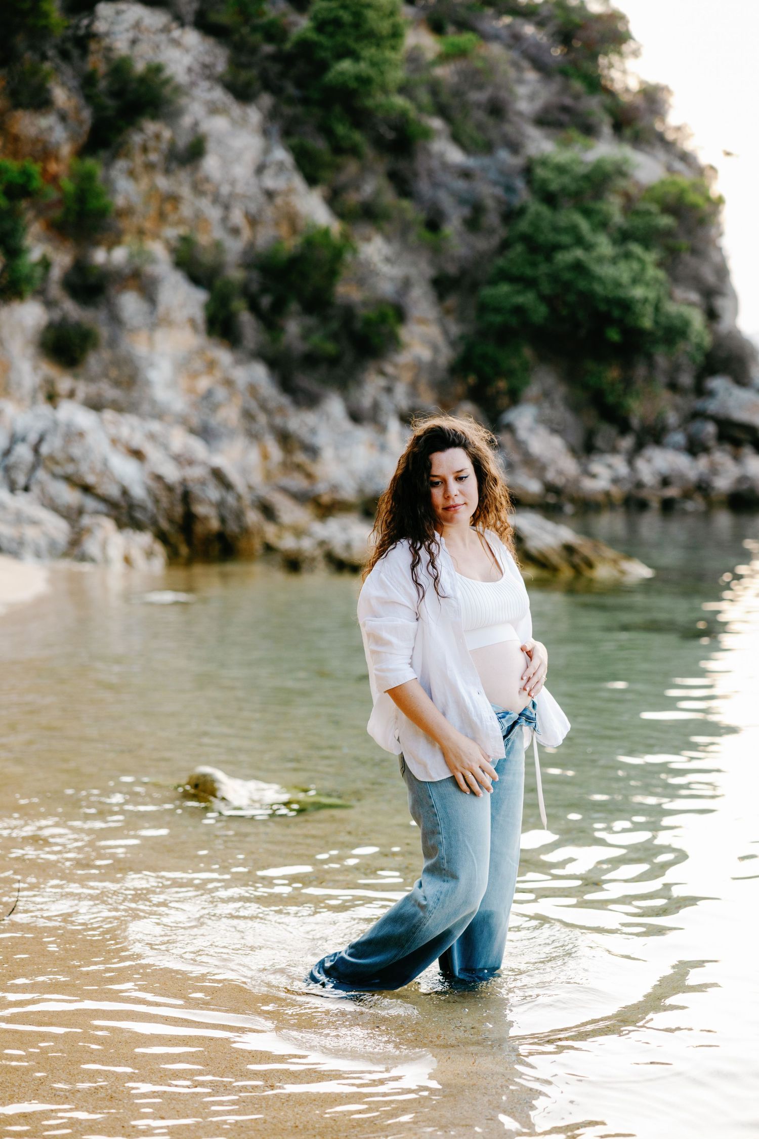 A woman in white shirt and jeans poses playfully by the water near rocky cliffs.