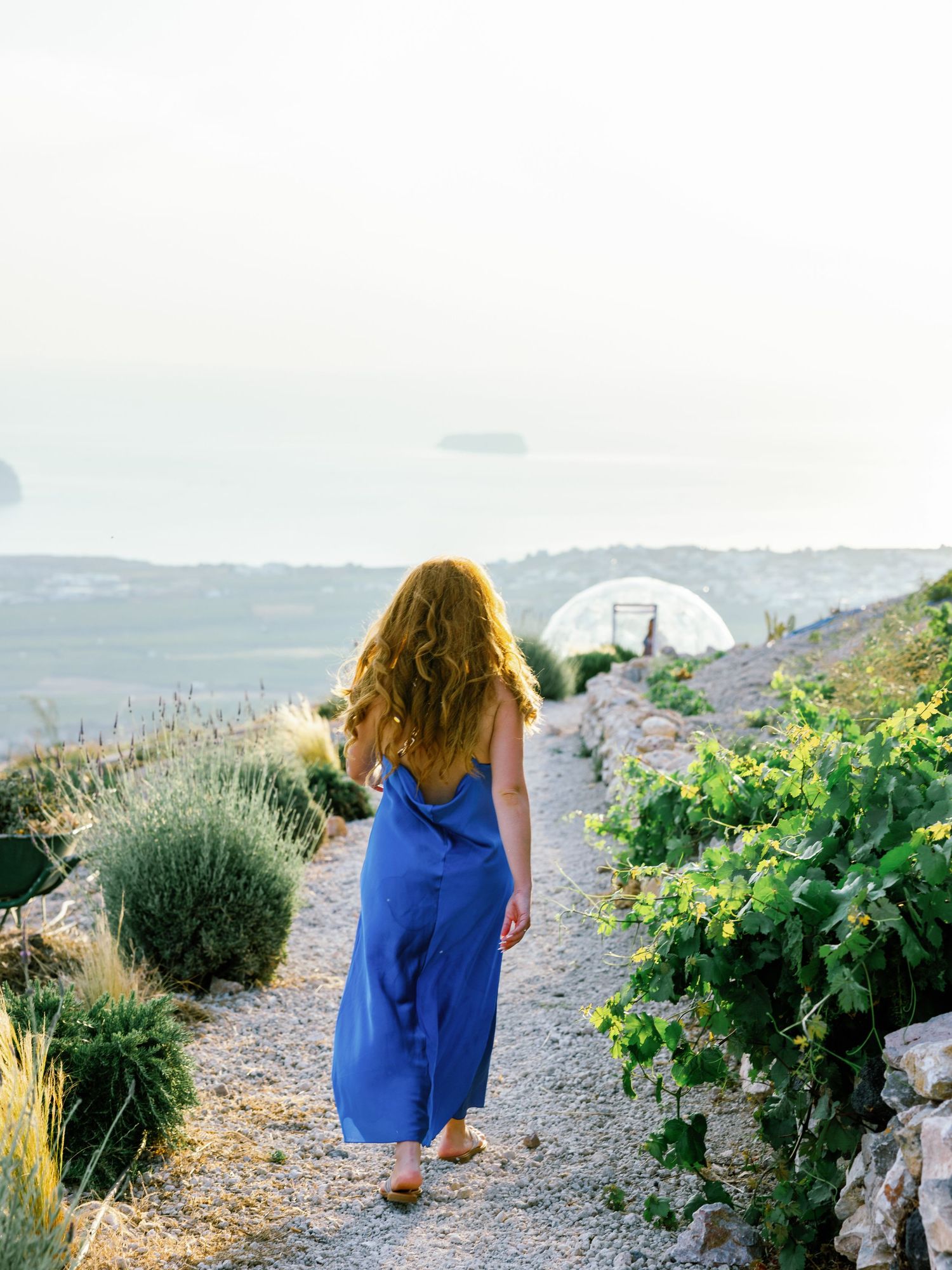 Woman in blue dress walking garden path with coastal mountains in background.