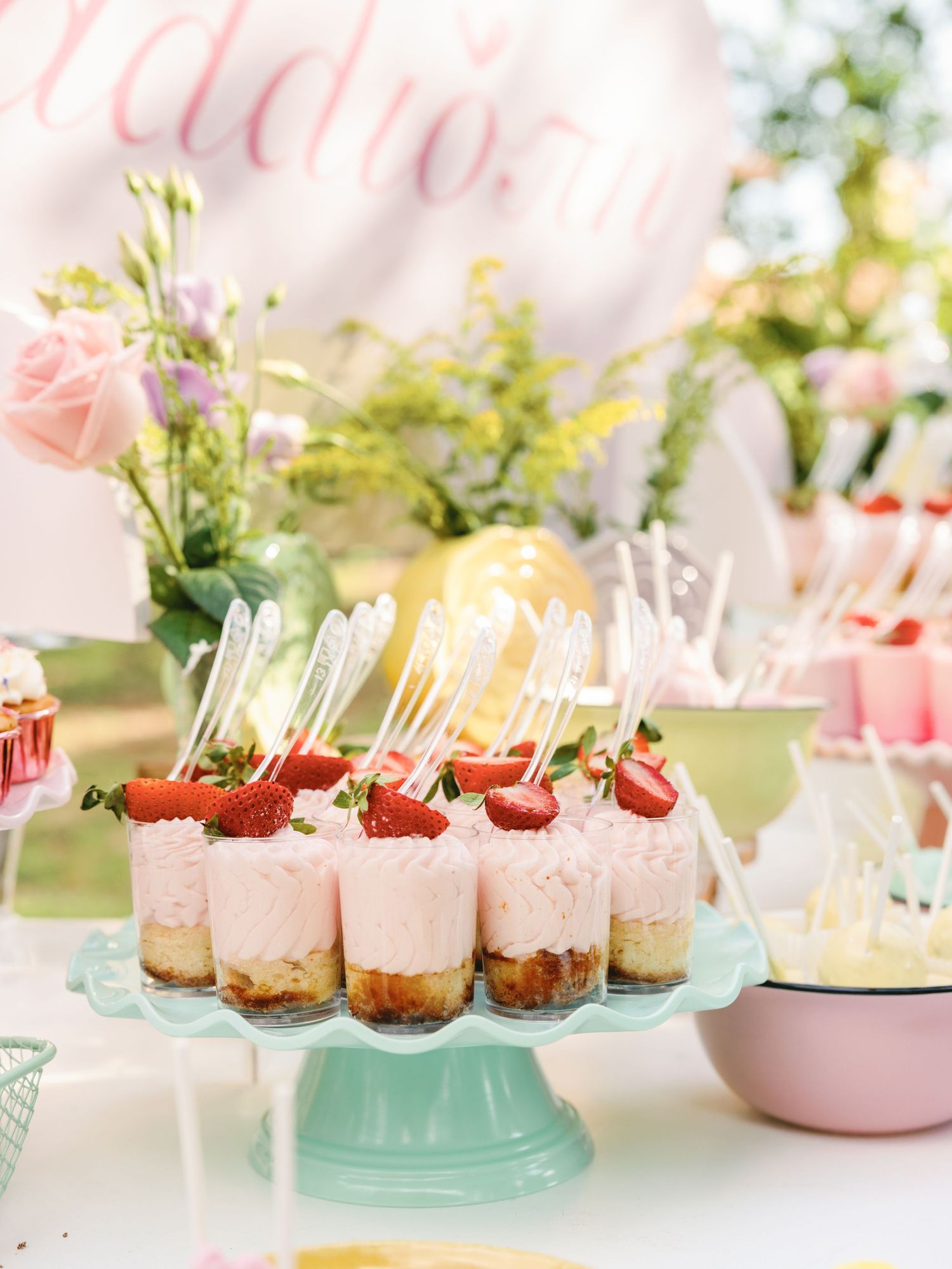 Mini desserts displayed on pastel pink and mint green cake stands with strawberry garnishes at an elegant party setup.