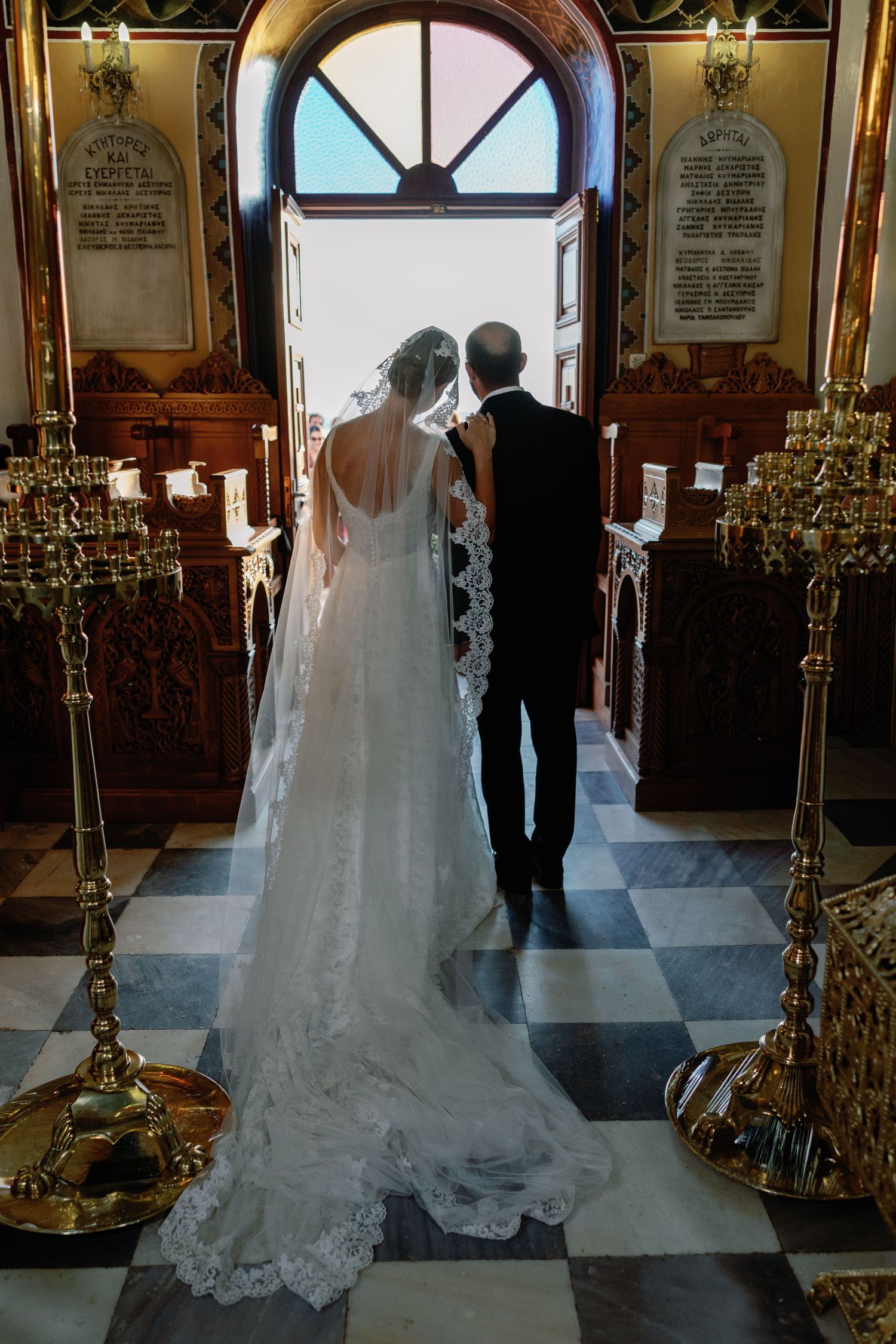 Bride and groom walking down church aisle toward open doors, bride in lace wedding gown with long veil, golden candlesticks.