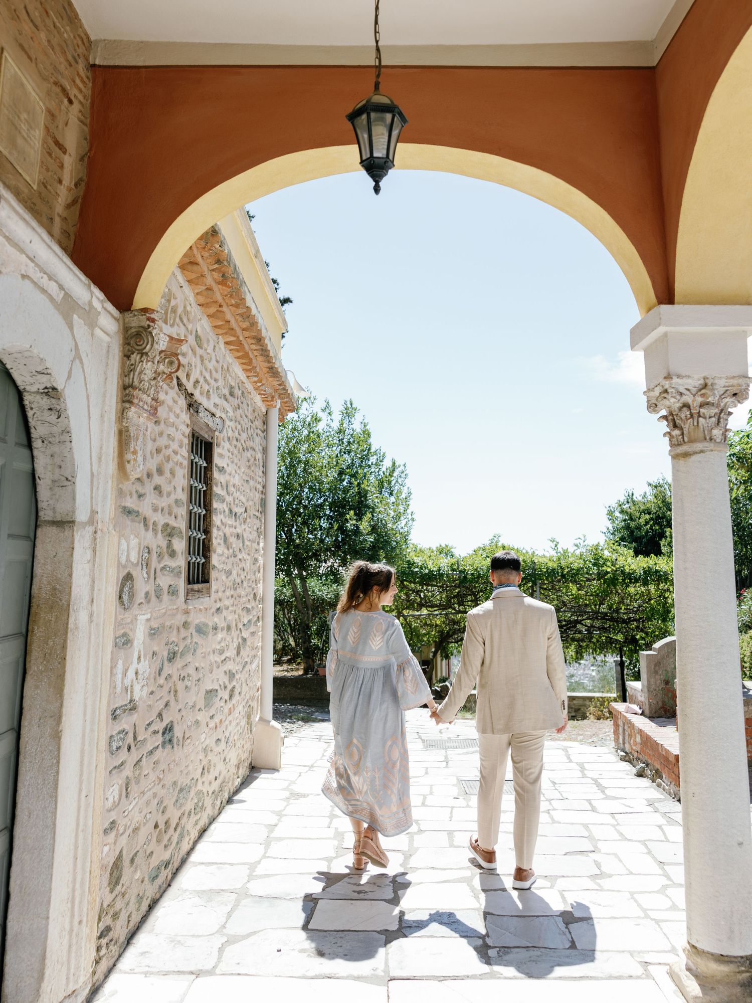 A couple holding hands while walking through a Mediterranean stone archway with hanging lantern and white columns.