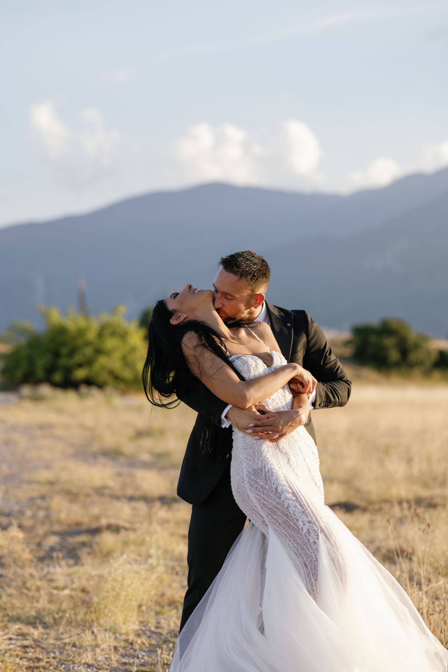 Romantic wedding couple embracing in a field with mountain backdrop at golden hour.