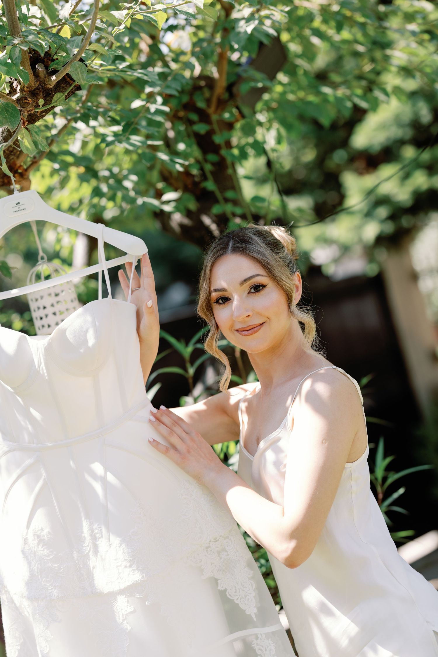 A bride in a white wedding dress poses under green foliage and sunlight.