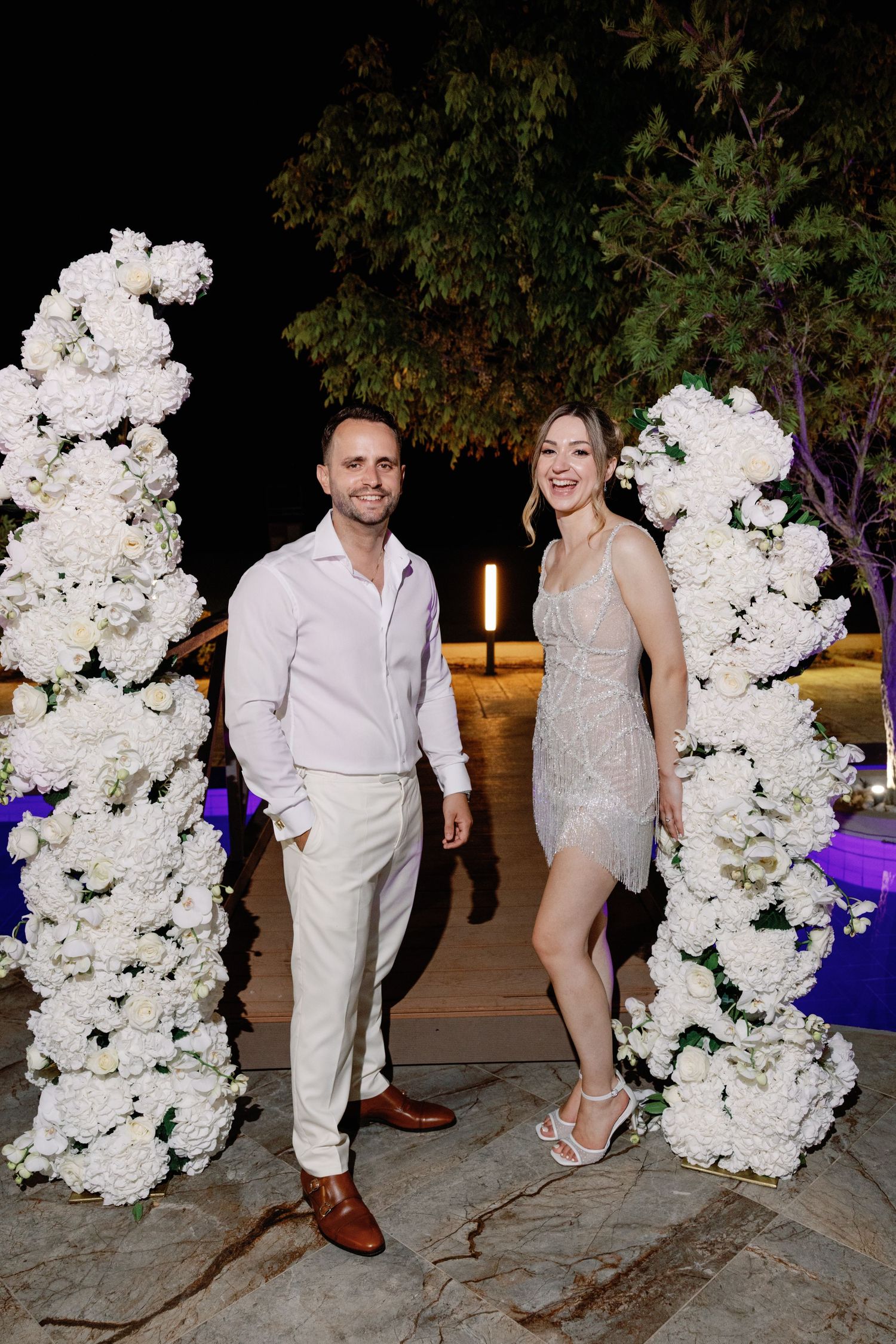 Wedding couple poses by elegant white floral arrangements at an evening reception.