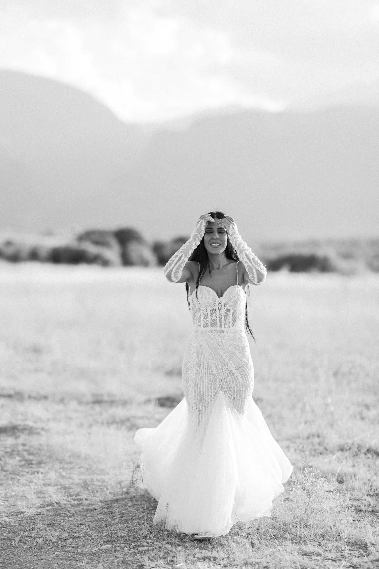 A bride in a flowing white dress dances in a desert landscape with mountains behind her.