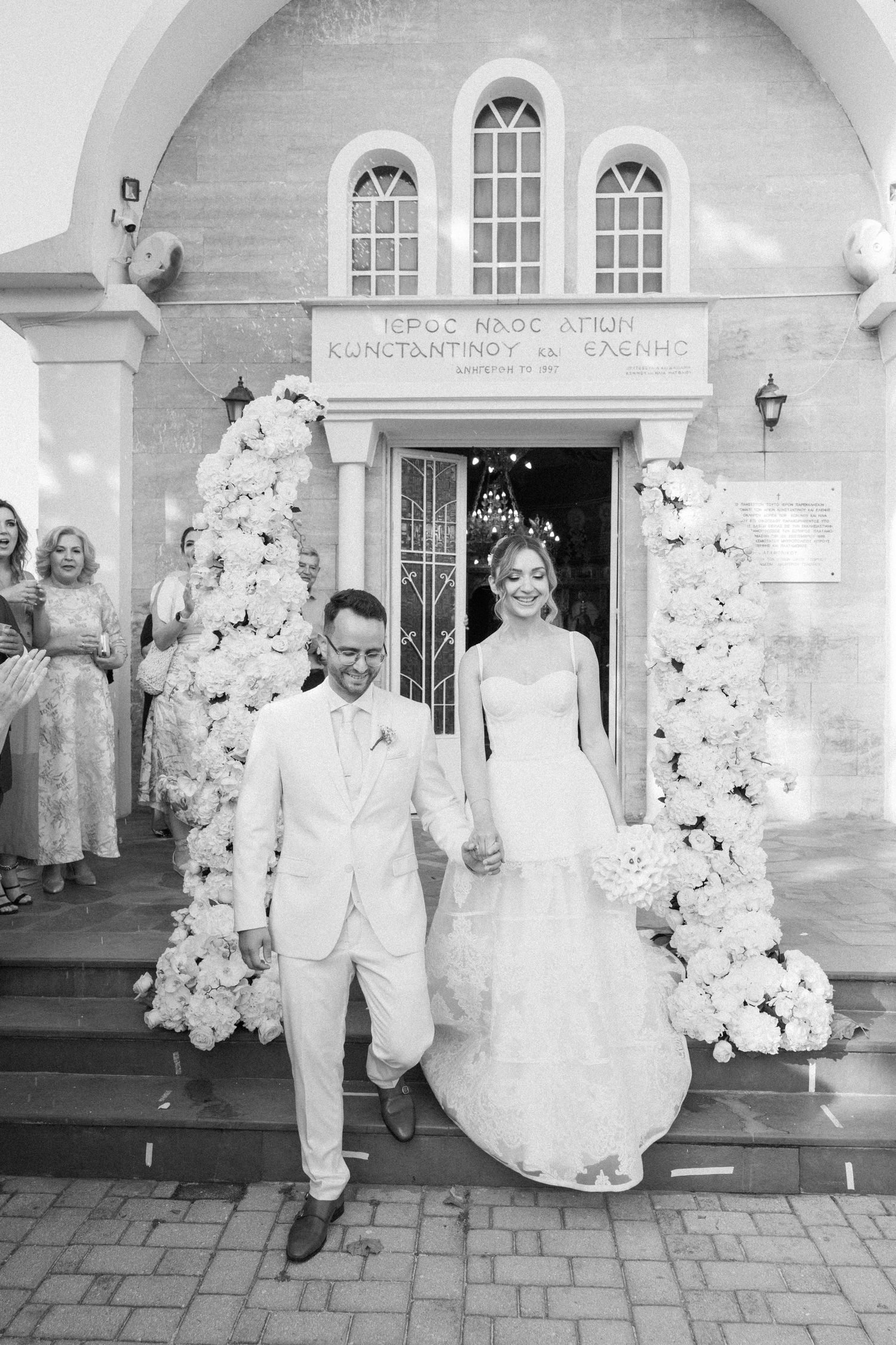 Newlyweds exiting white church building decorated with elegant floral arrangements.