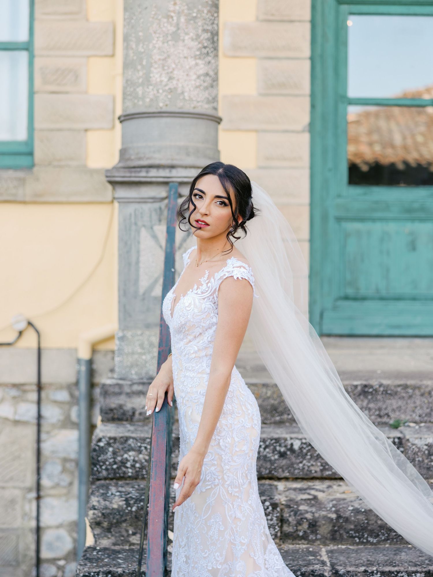 Bride in fitted lace gown with flowing veil poses against classical architecture.