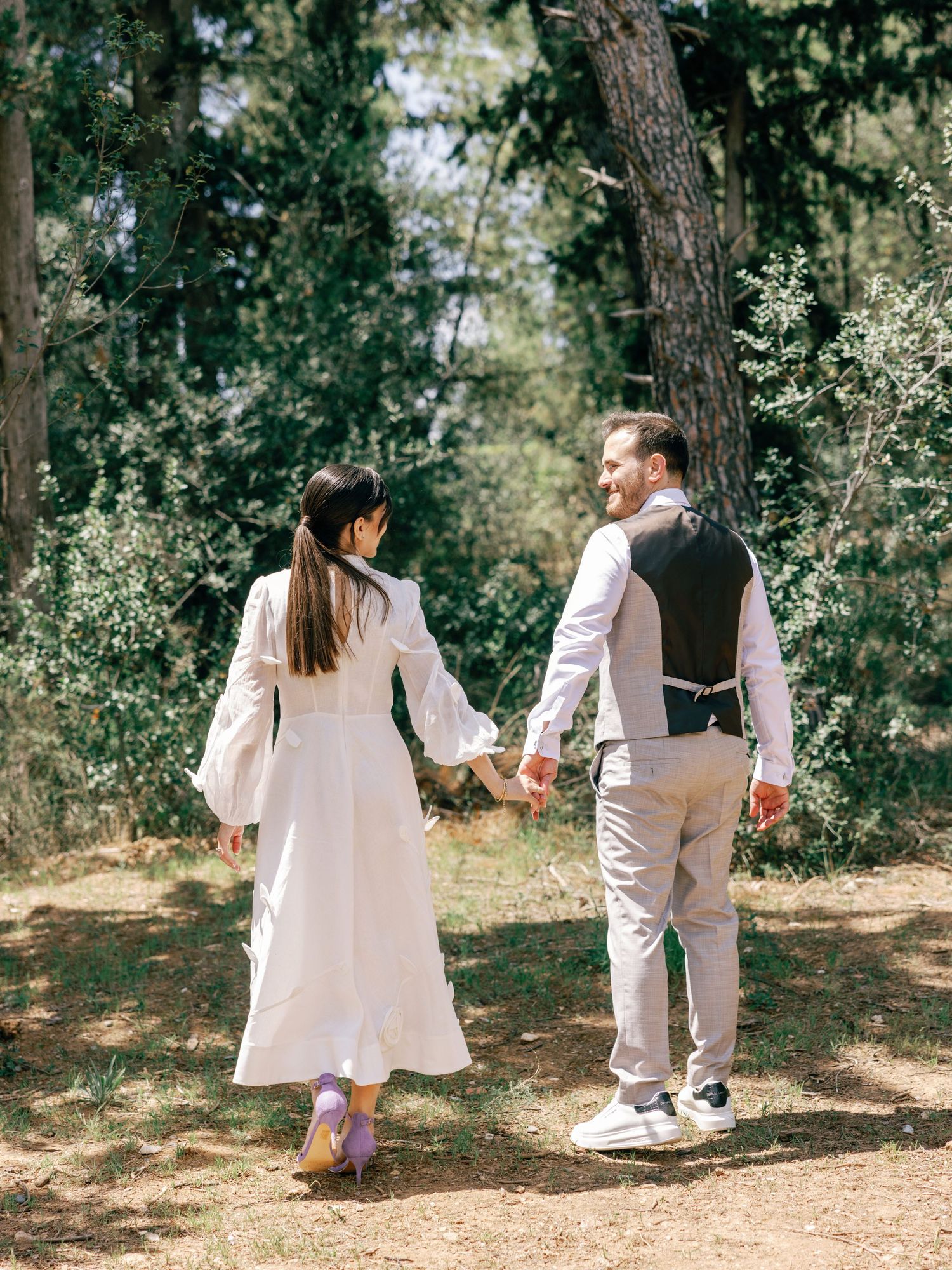 A couple in light-colored outfits holding hands while walking along a forest path on a sunny day.