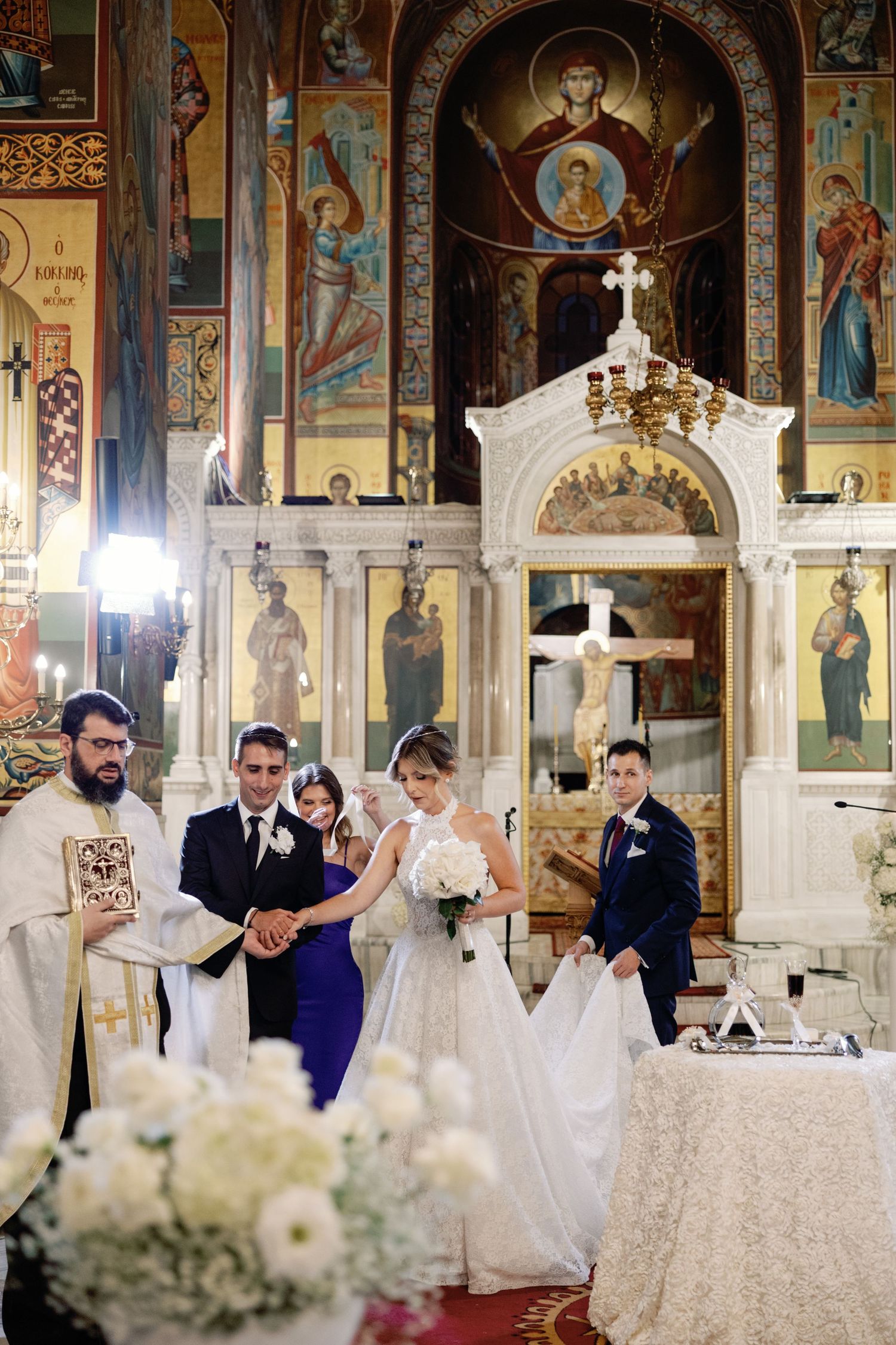 Orthodox wedding ceremony taking place in an ornate church with beautiful religious frescoes and white floral decorations.