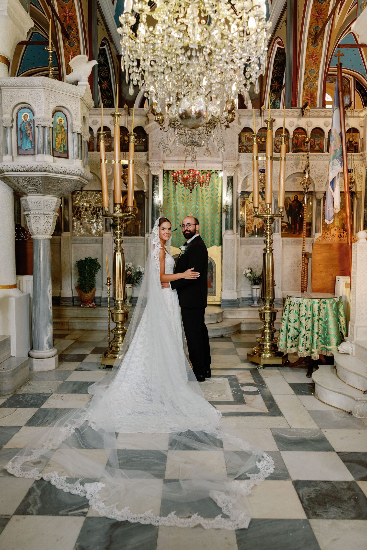 Bride and groom pose in ornate Orthodox church with chandelier, checkered marble floor, and golden iconostasis.