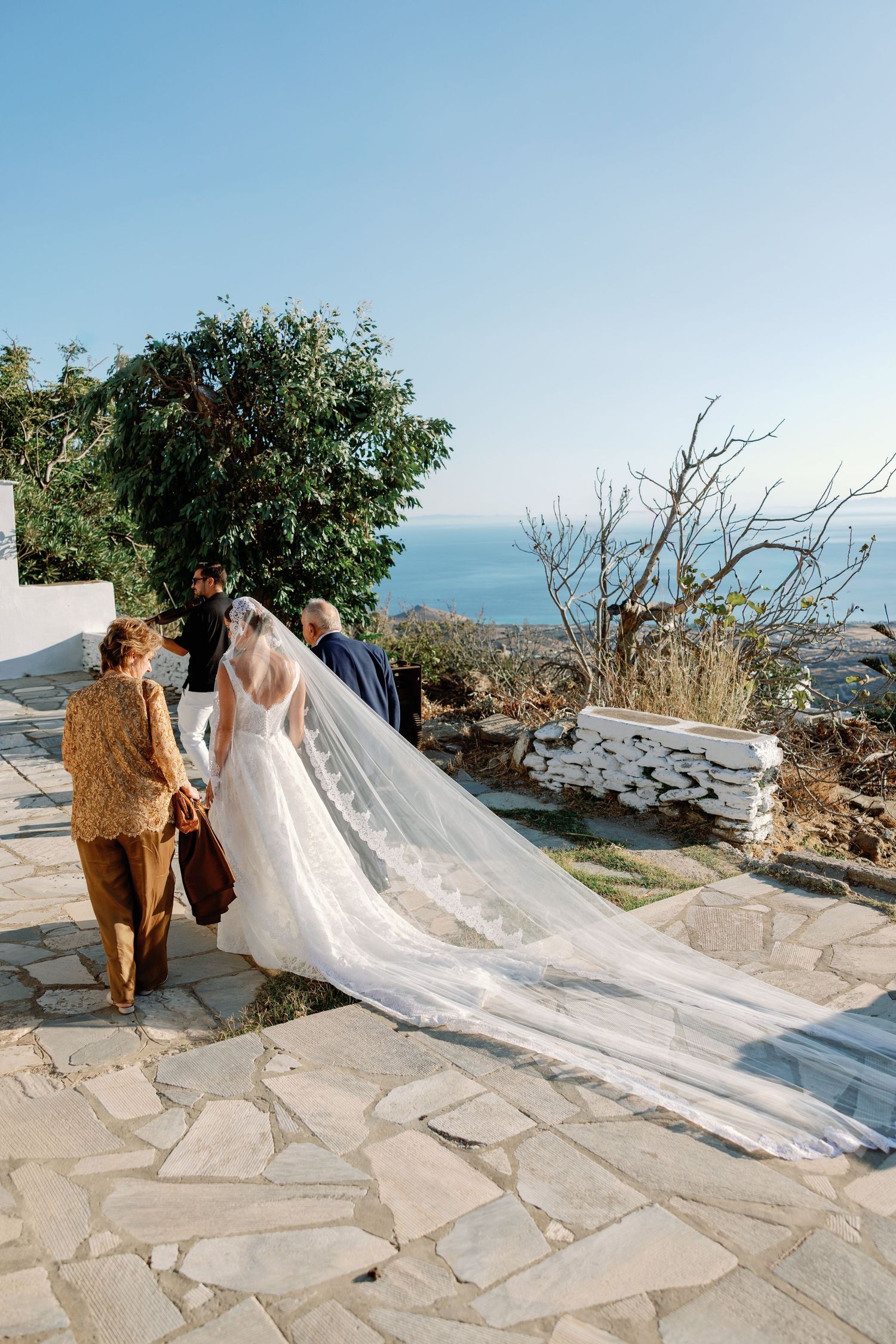 Bride in lace gown with long veil walks with guests on stone terrace overlooking the sea at a Greek island wedding.