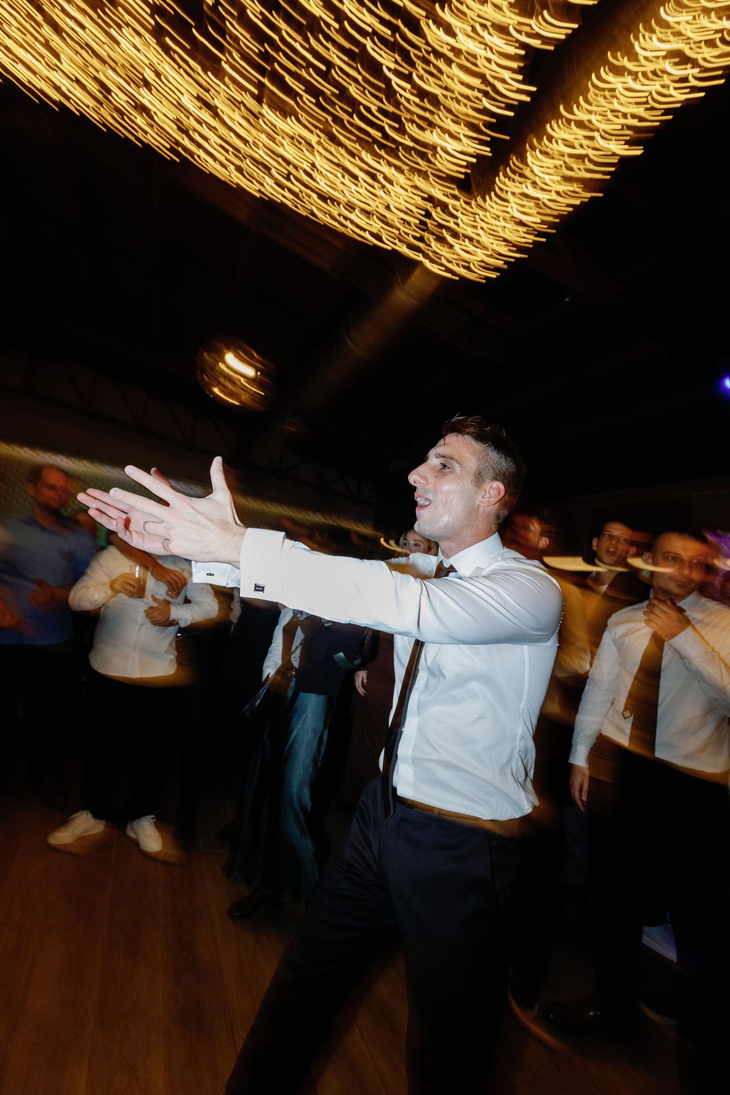 Wedding guests perform a synchronized dance routine under decorative ceiling lights at a reception.