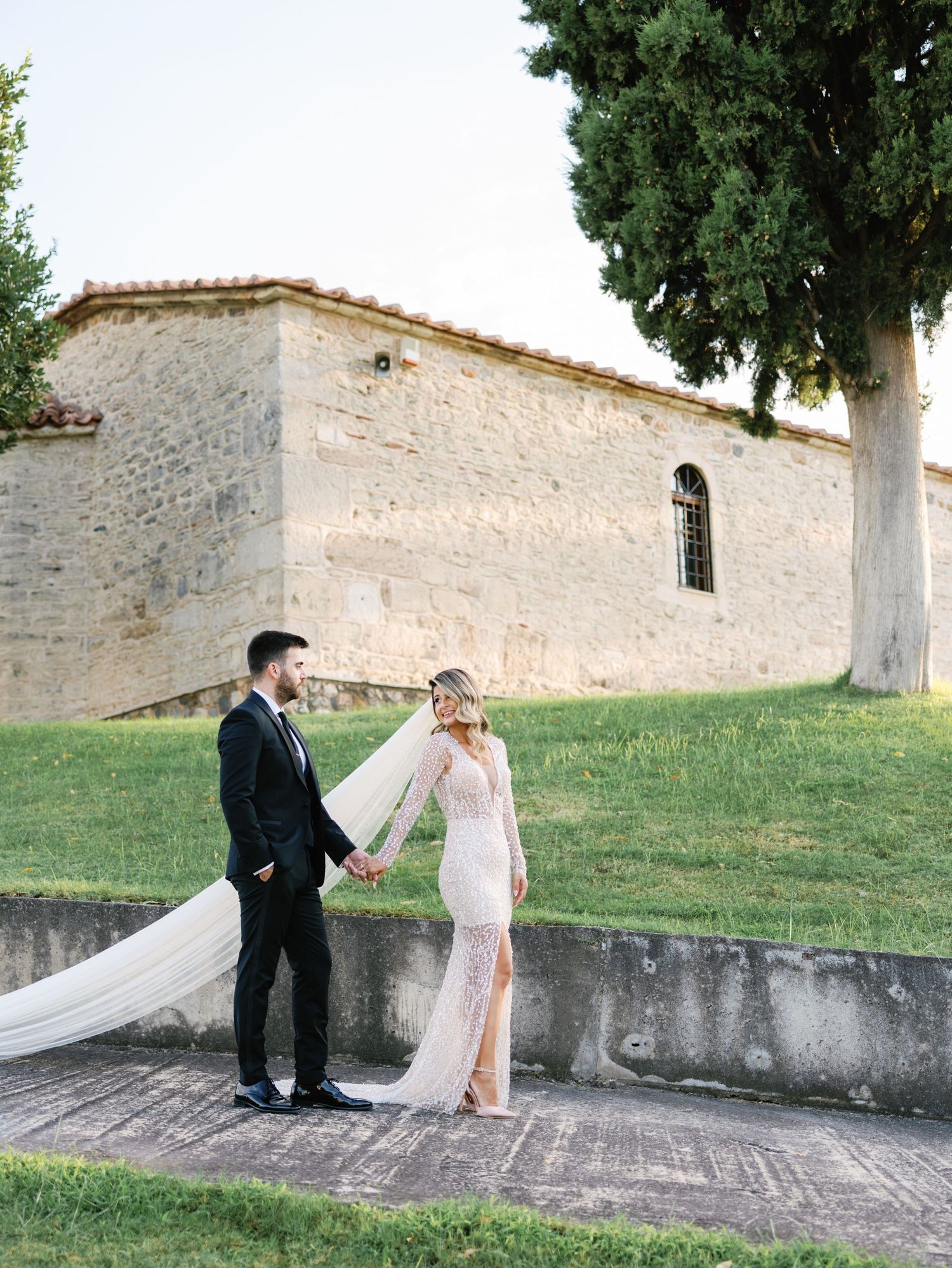 A couple poses together outside a rustic stone church with cypress trees in an elegant wedding photo.