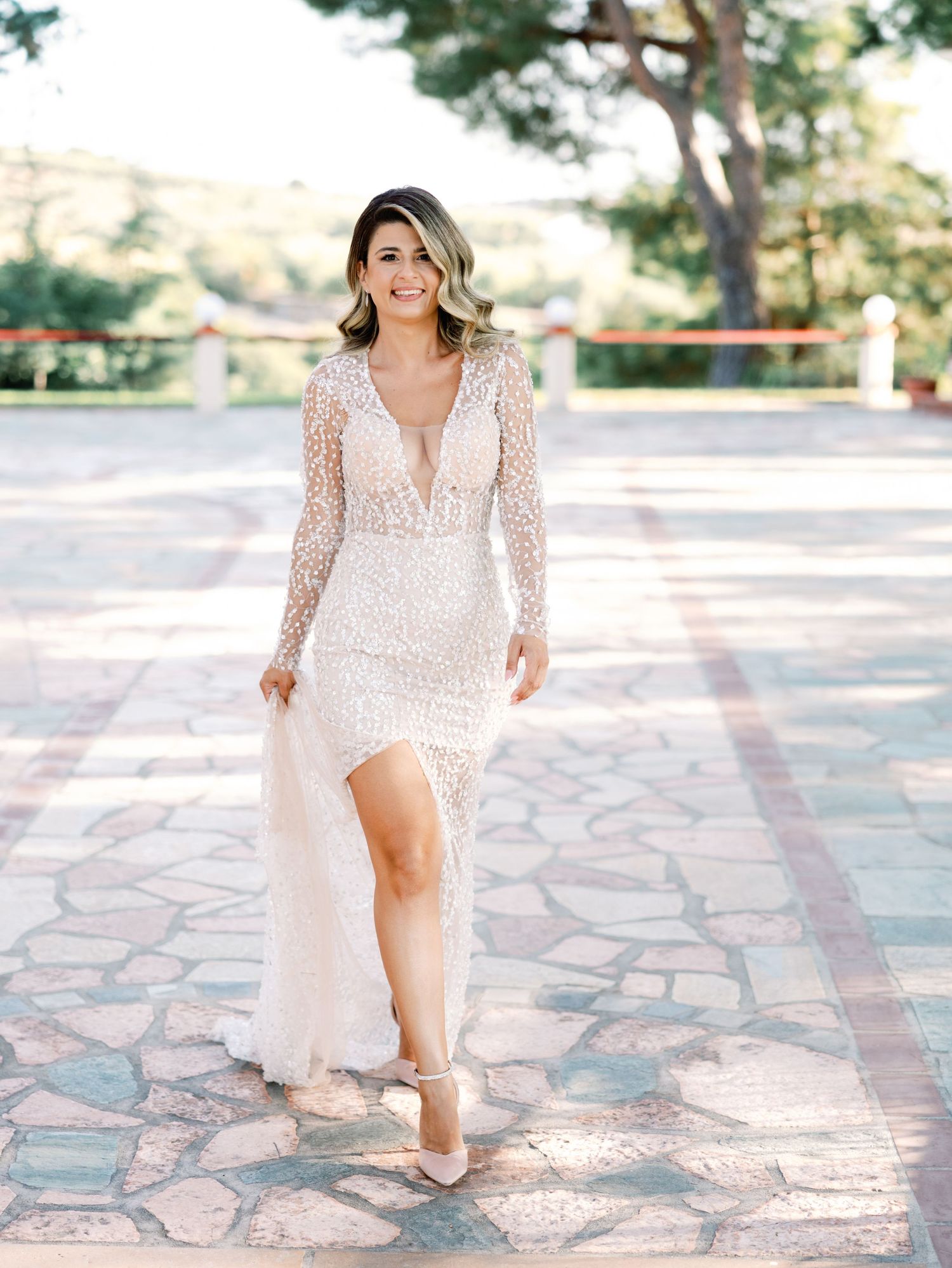 Elegant bride in beaded gown poses on stone patio during sunlit outdoor photo session.