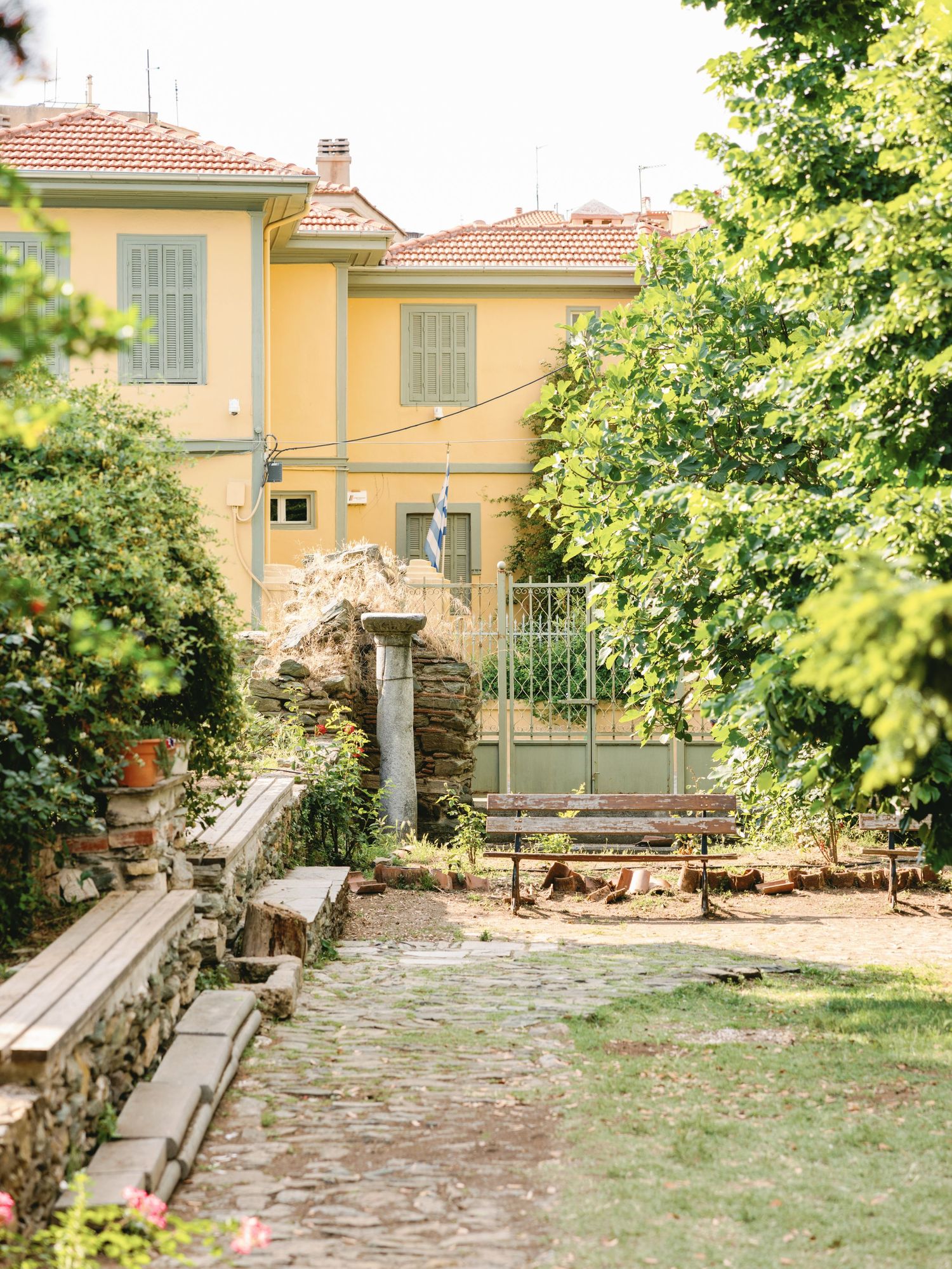 Charming yellow Mediterranean villa with stone pathway and garden benches surrounded by lush green foliage.