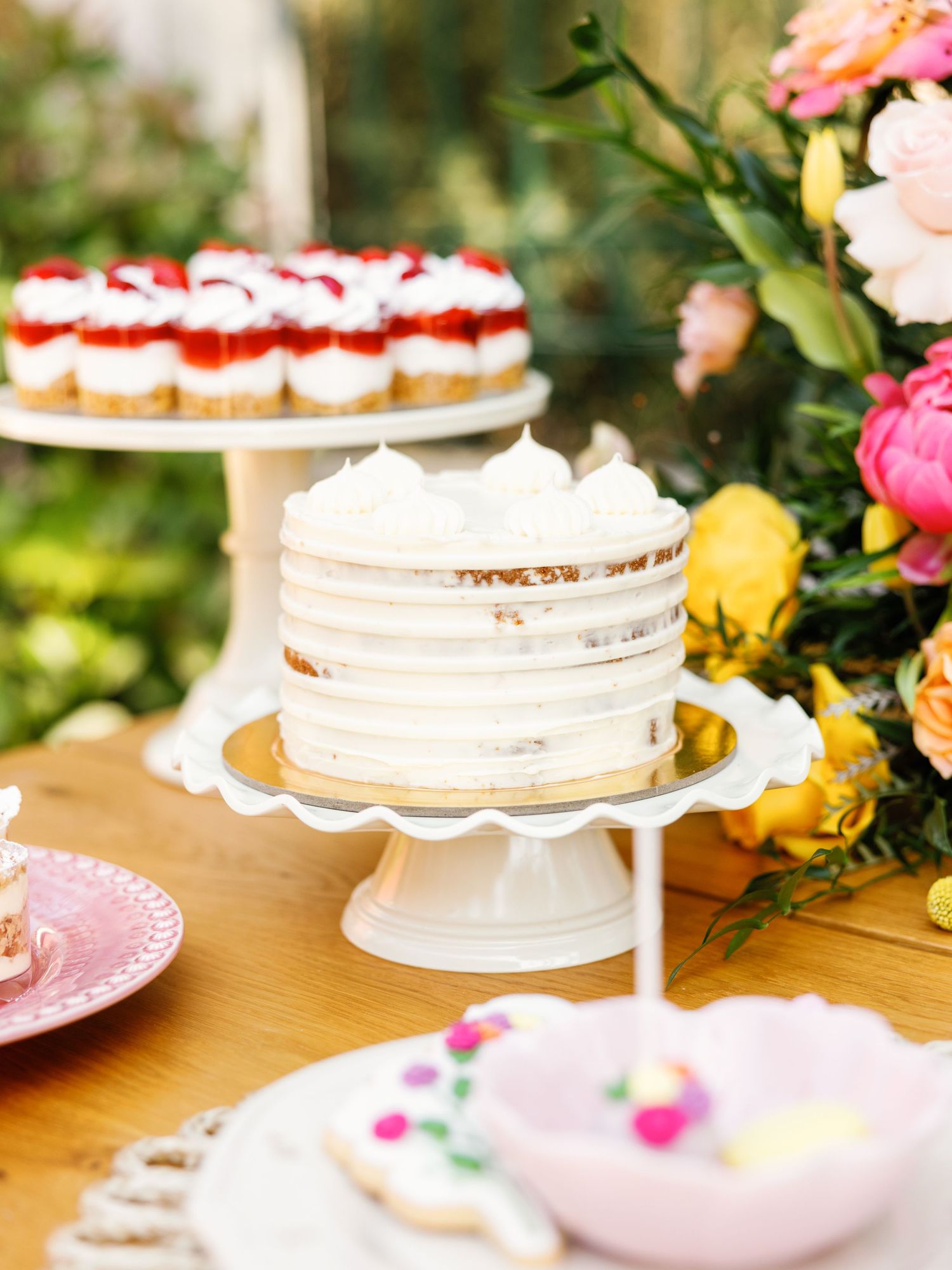 White frosted layer cakes on decorative pedestals surrounded by pink and yellow flowers on a wooden table.