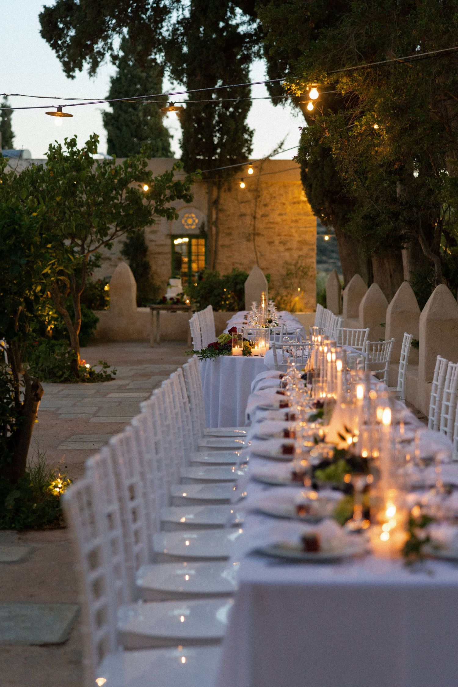 Elegant outdoor wedding reception with long white tables, candles, and string lights at dusk.