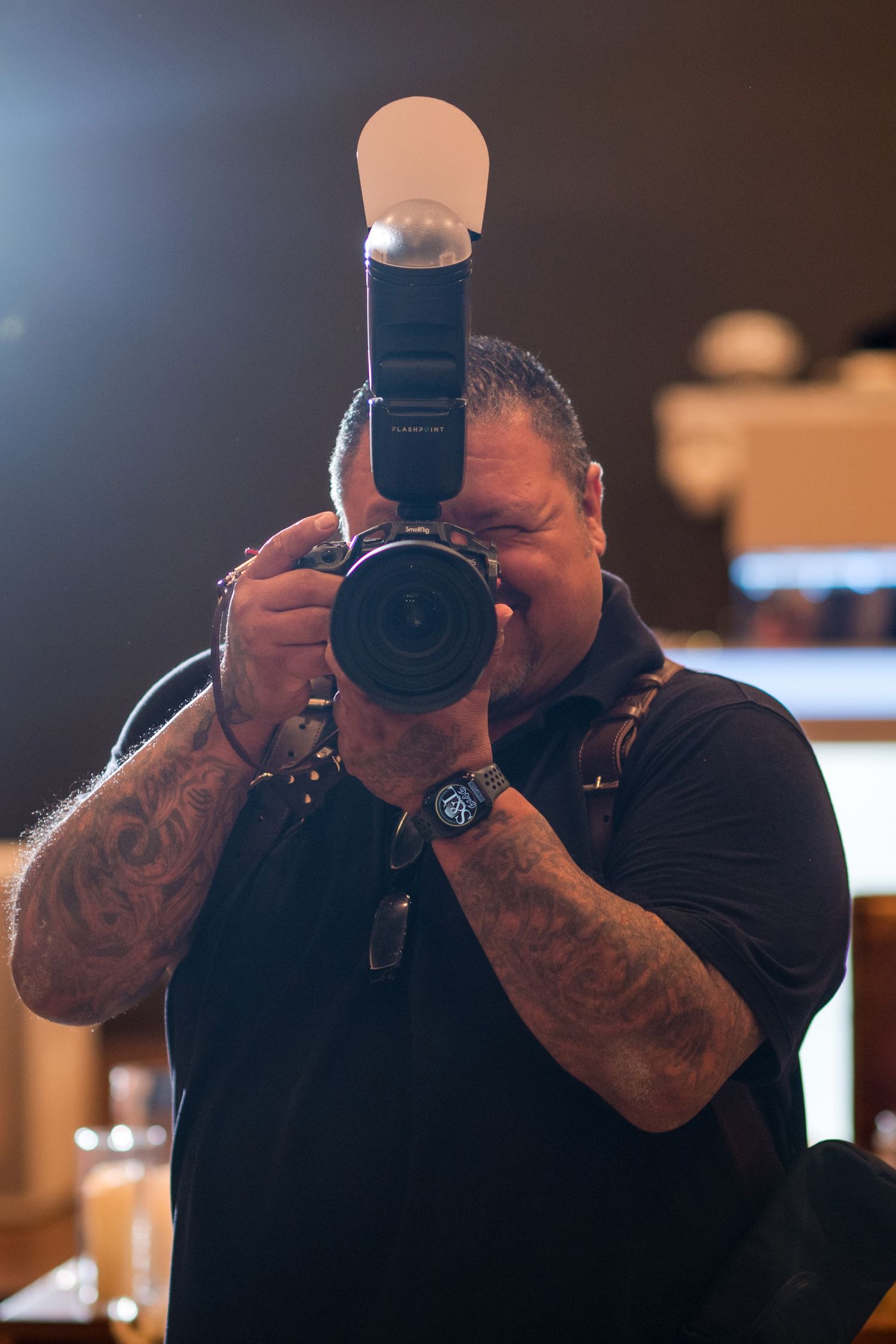 Wedding photographer with tattoos holds professional camera with flash diffuser in indoor venue.