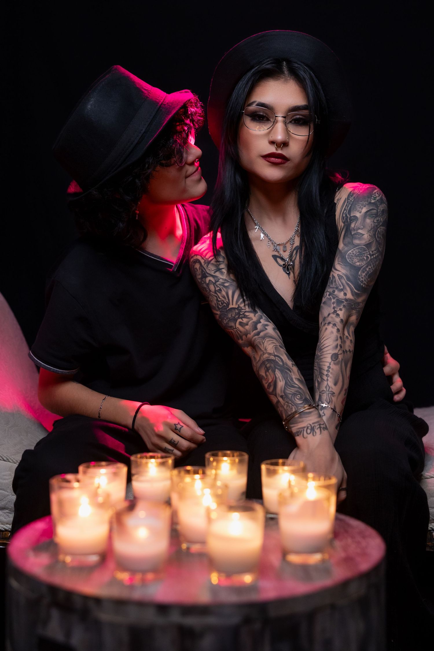 Two people in black outfits and hats pose dramatically with lit candles creating moody red lighting.