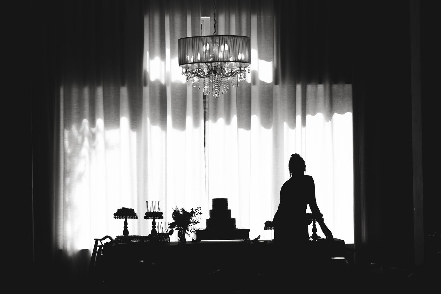A silhouetted figure sits at a table against white curtains with a chandelier hanging above in a dramatic black and white scene.