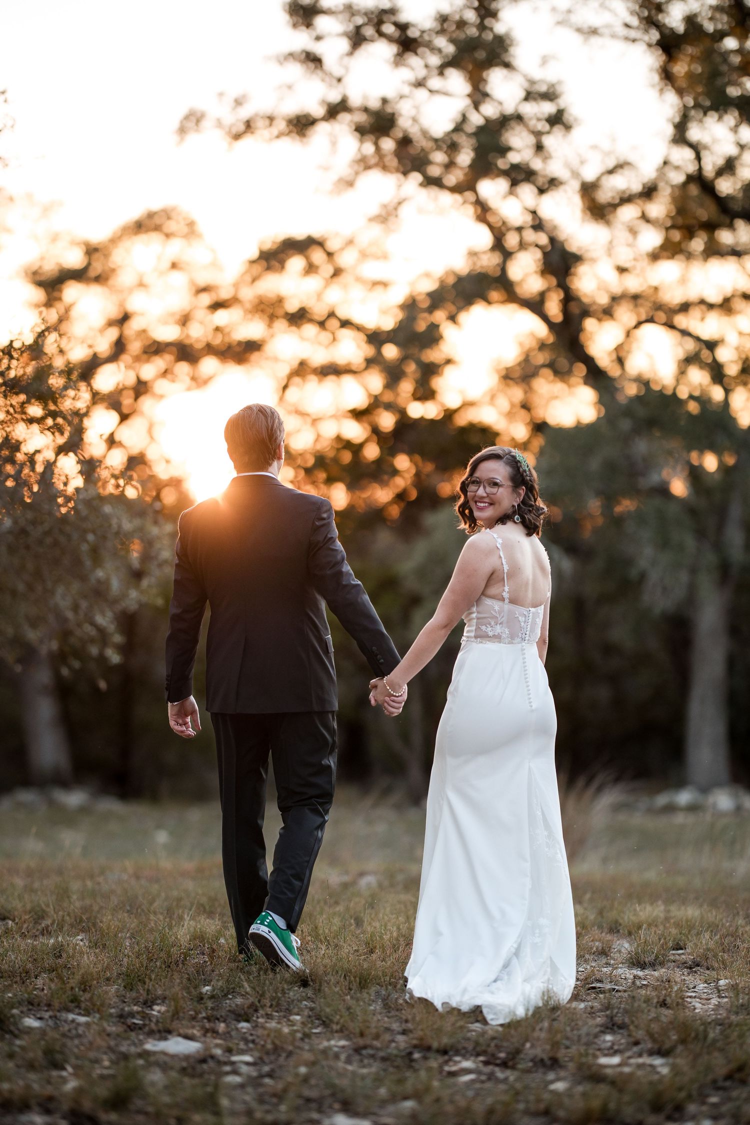 A couple in wedding attire walks hand in hand through a field at sunset with golden light filtering through trees.