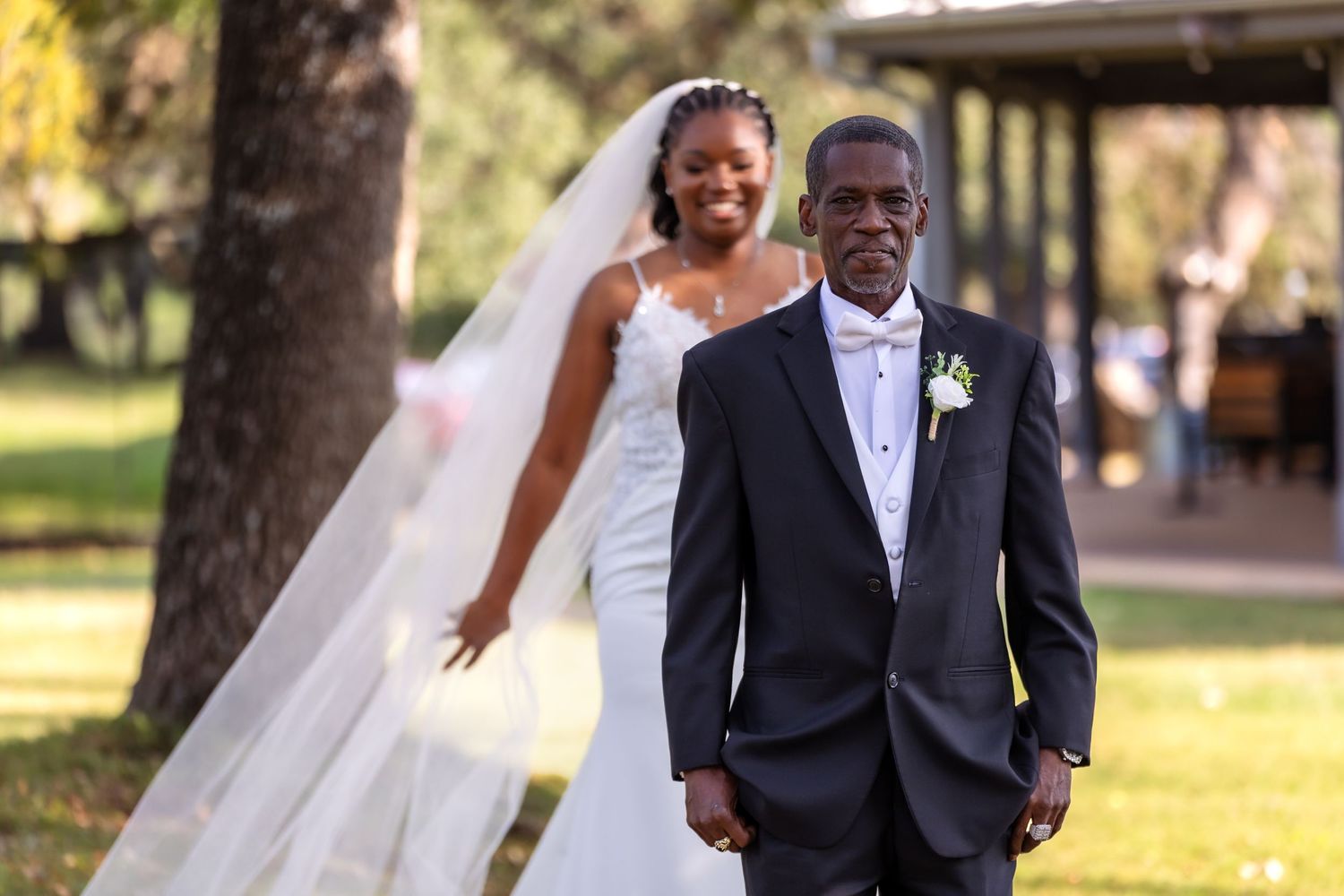 A happy couple on their wedding day standing outdoors in formal attire with a flowing white veil and boutonniere.