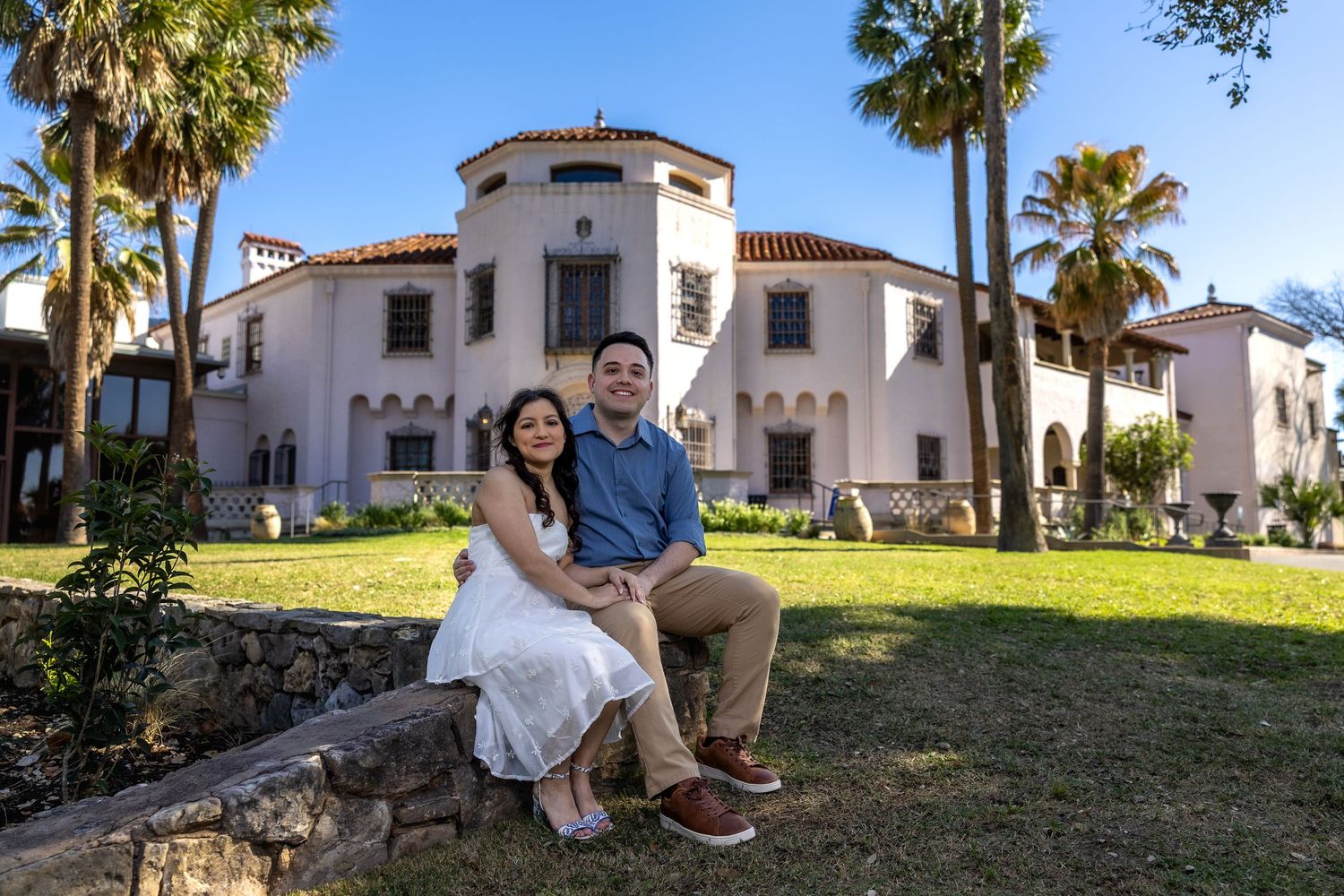A couple sits together on a stone bench in front of a Spanish-style white stucco building with palm trees.