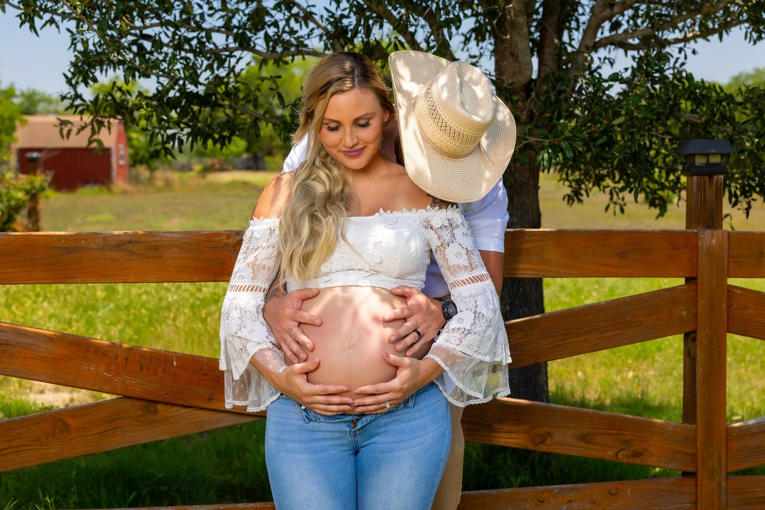 Maternity photo of an expectant mother in white lace and jeans standing by a wooden fence in a sunny field.