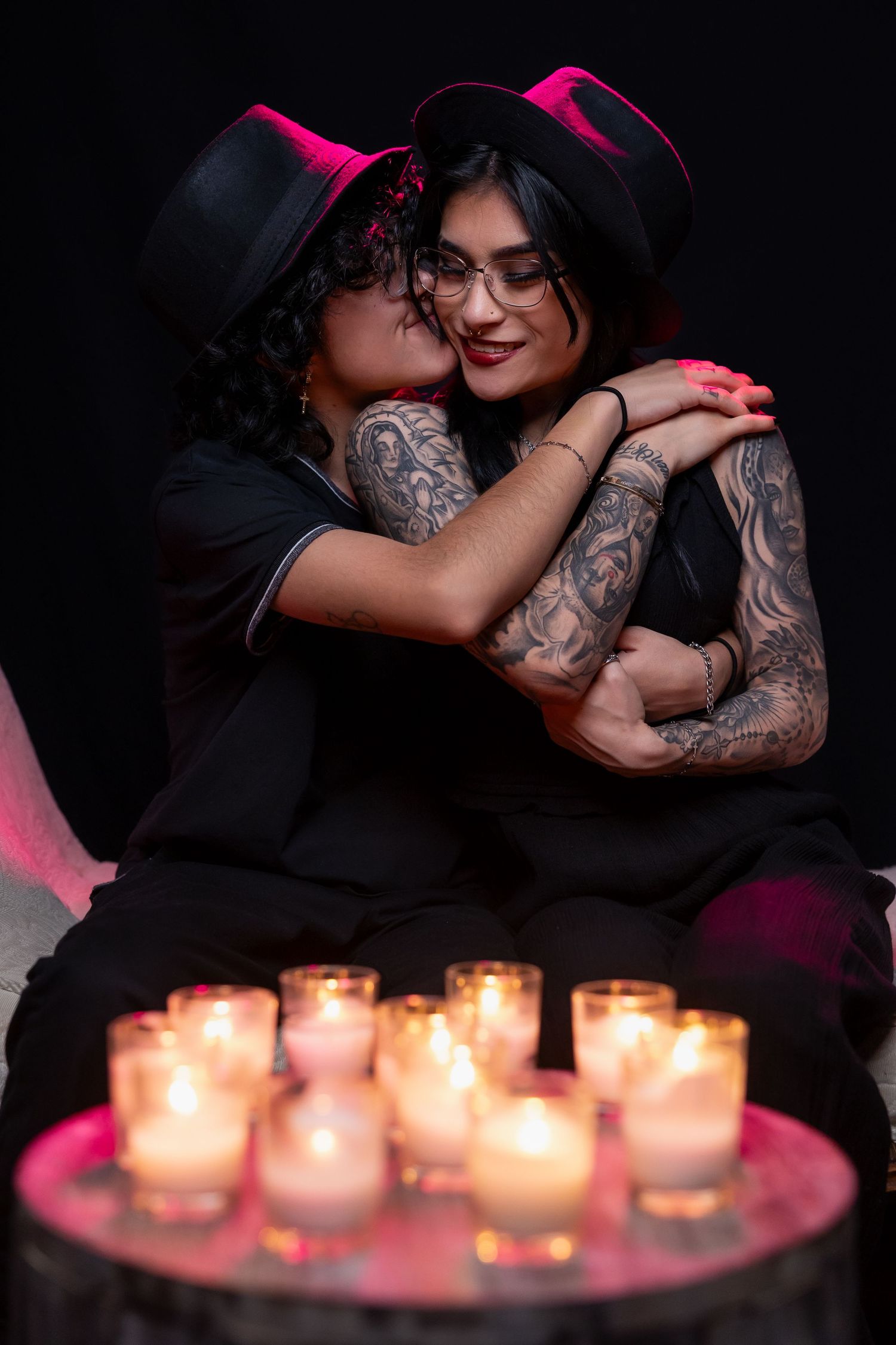 Intimate portrait with candlelight featuring two people in black attire and hats sharing an embrace.