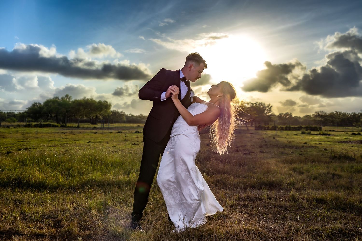 A bride and groom share a romantic dance in a sunlit field at sunset, with dramatic clouds in the background.