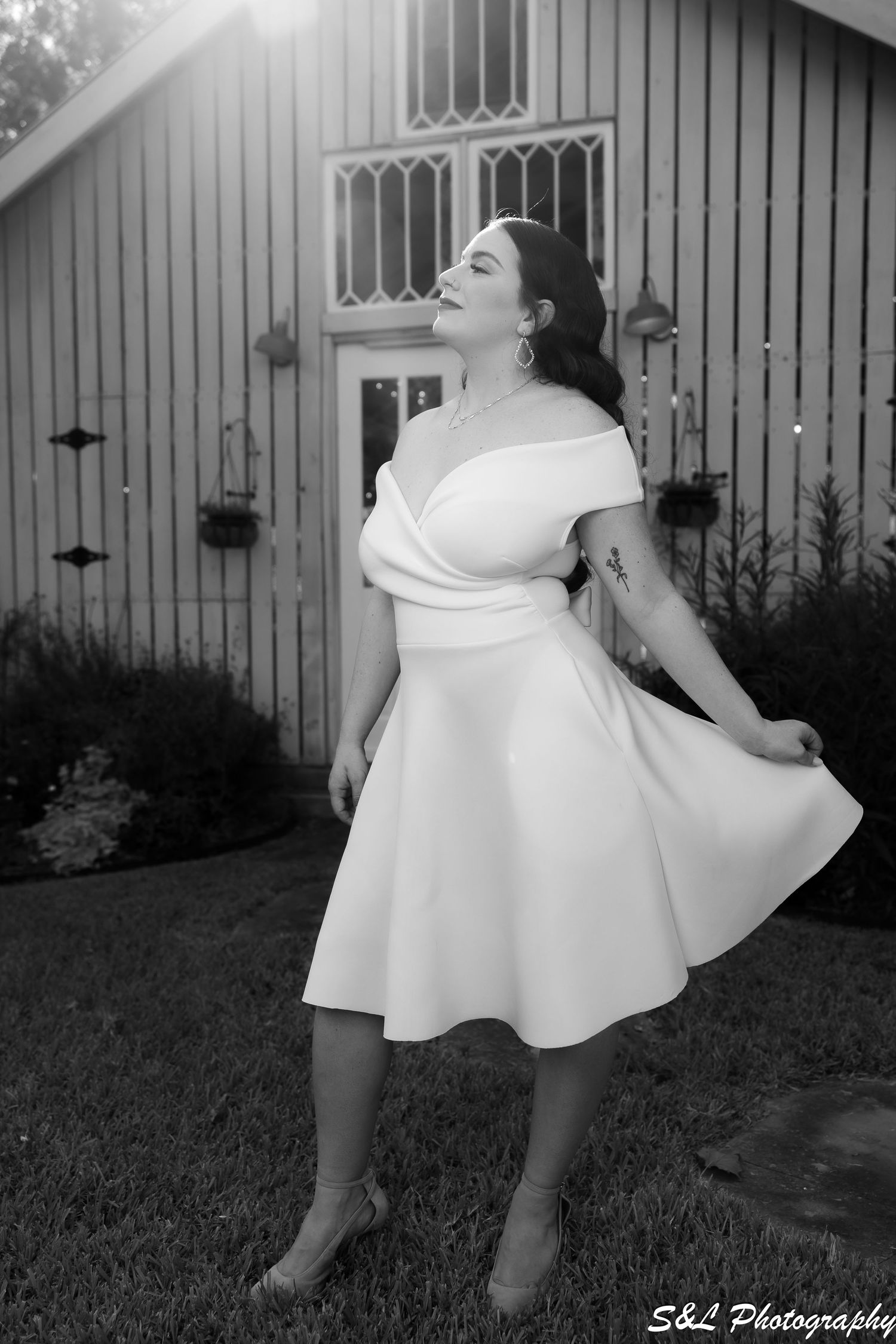 Black and white portrait of someone in a white dress twirling in front of a rustic barn.