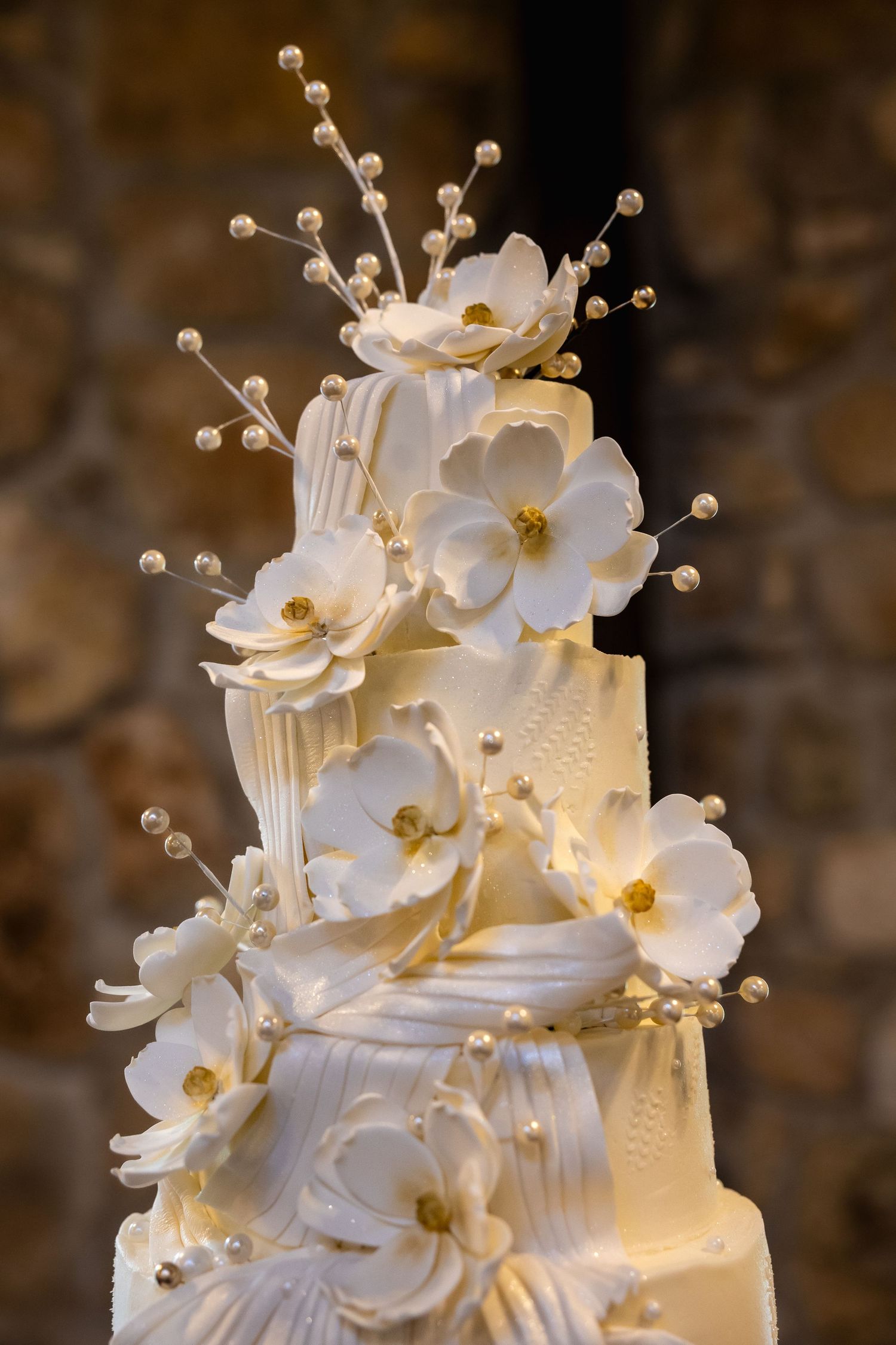 Elegant white wedding cake decorated with delicate sugar flowers and pearl accents against a textured brown background.