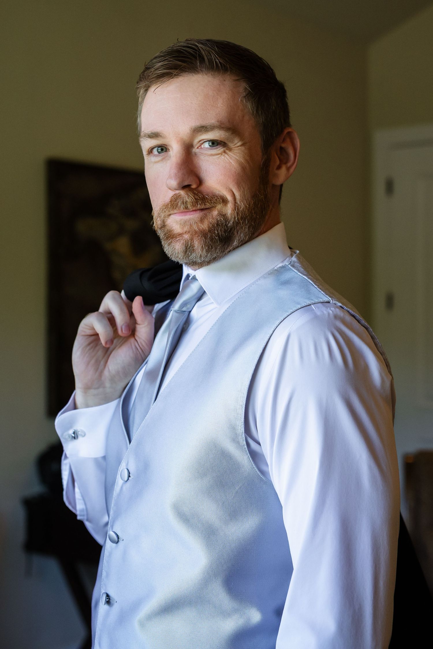 A man in a light blue dress shirt and vest smiles while adjusting his collar in indoor lighting.