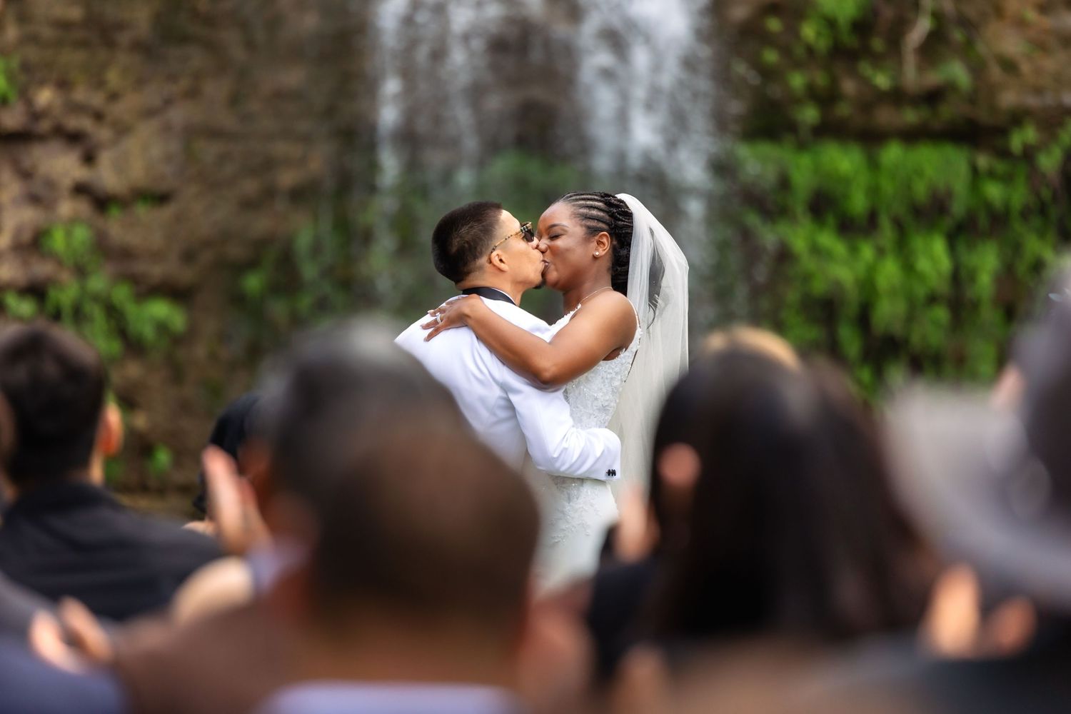 A couple shares a romantic kiss during their outdoor waterfall wedding ceremony as guests look on from the foreground.