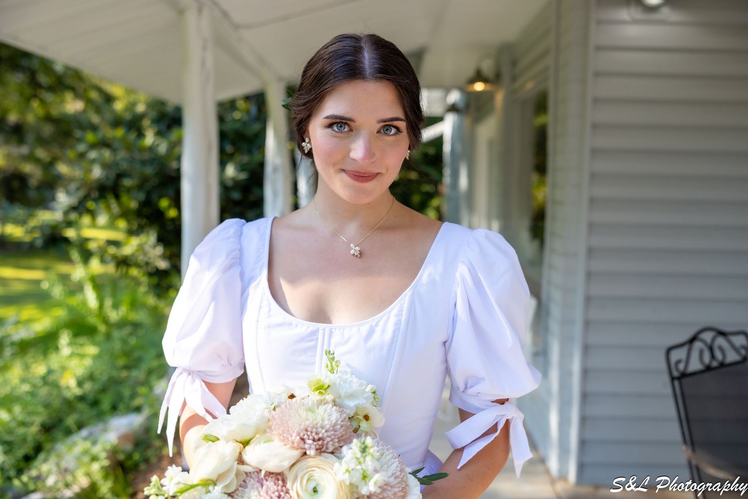 Portrait of someone in a white dress with puff sleeves holding a pastel floral bouquet on a porch.