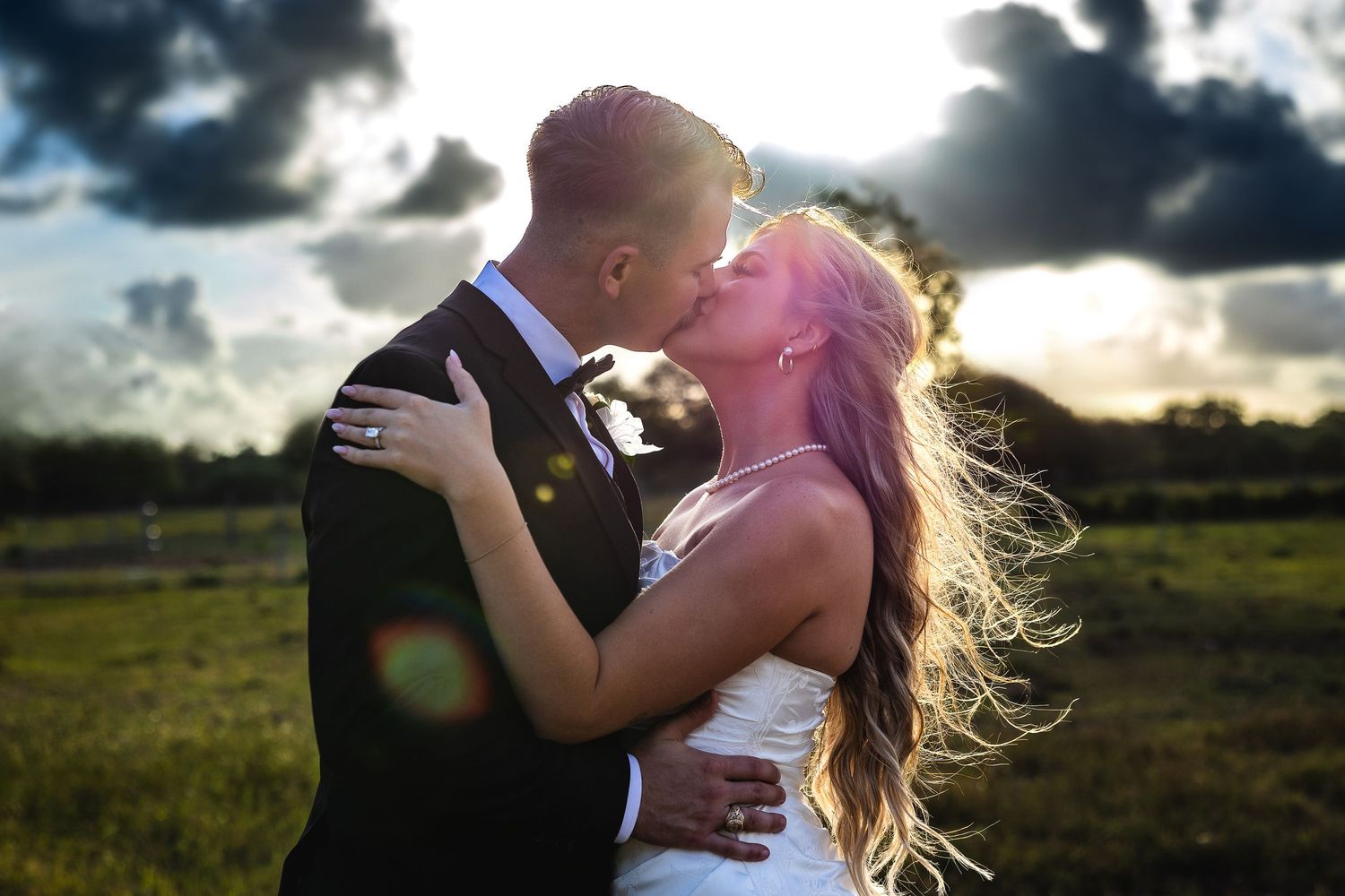 A bride and groom share a romantic kiss outdoors at golden hour with dramatic clouds in the background.