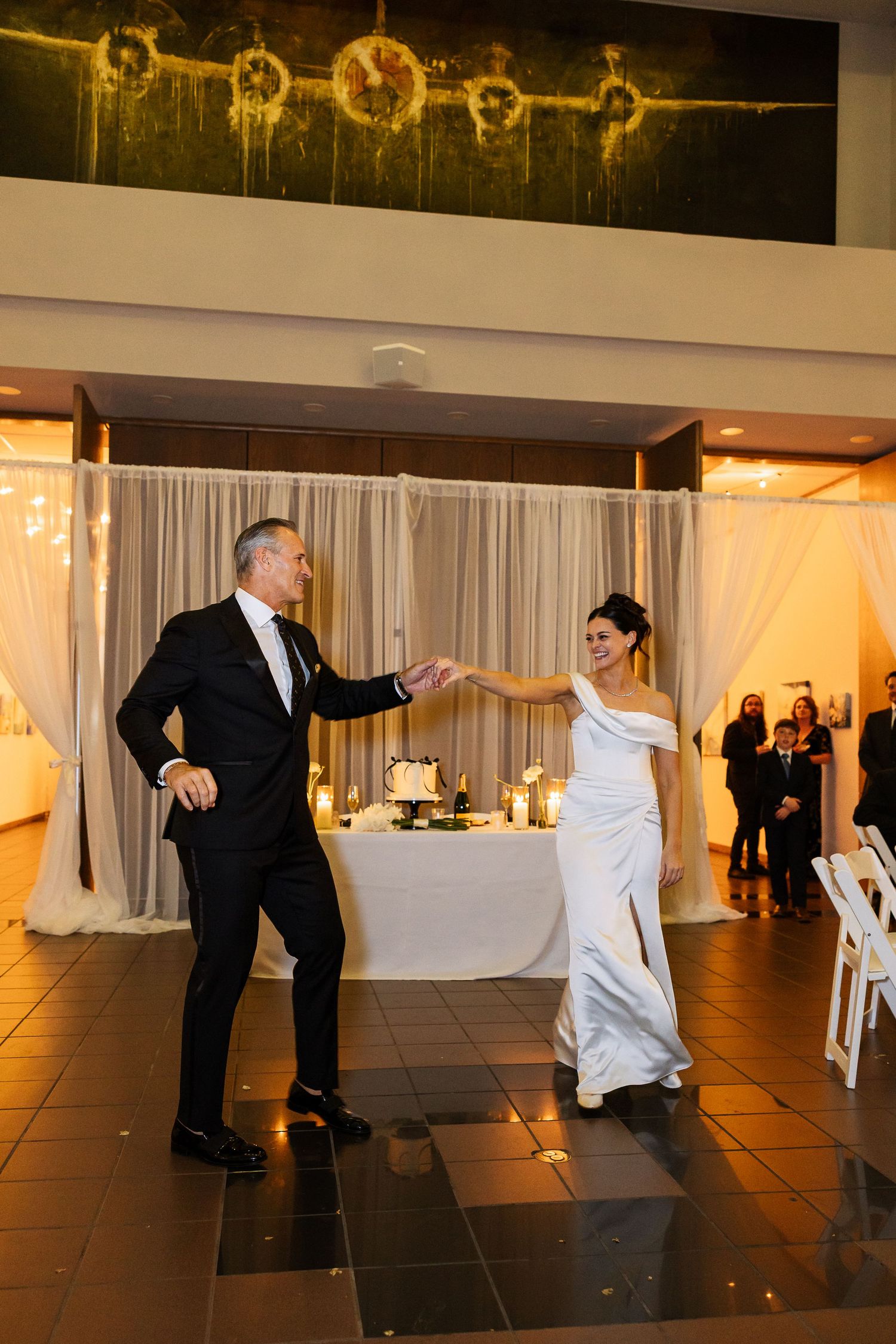 Wedding guests dance together on a dark tile floor with white curtains and ambient lighting in the background.