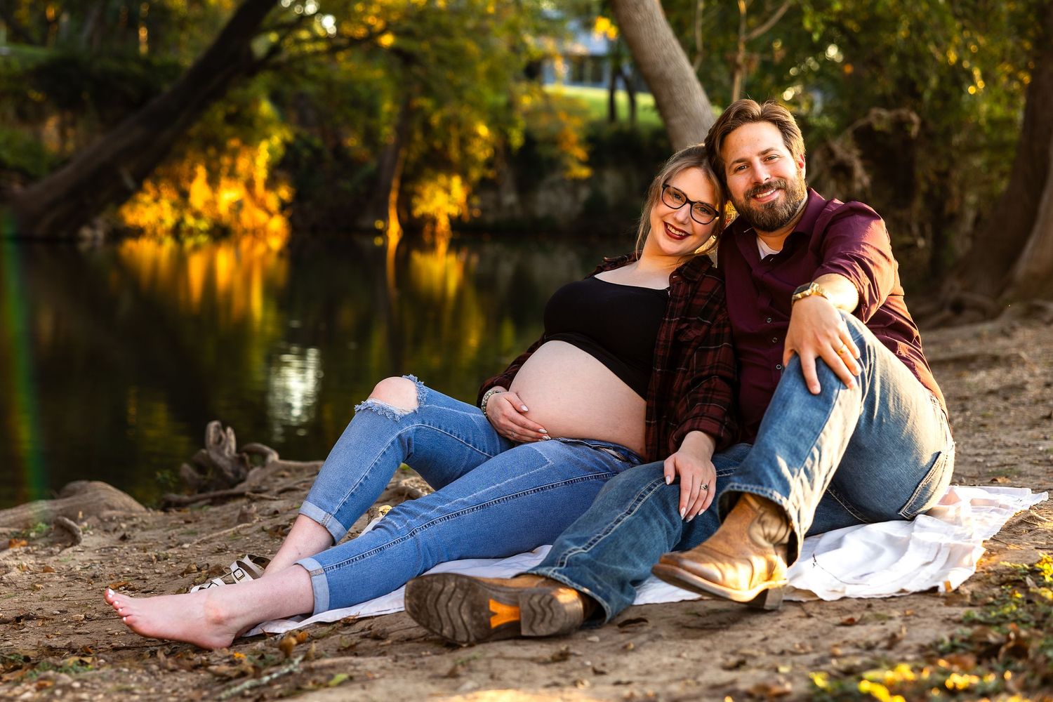A couple relaxes on a blanket by a scenic pond during autumn with fall foliage reflecting in the water.