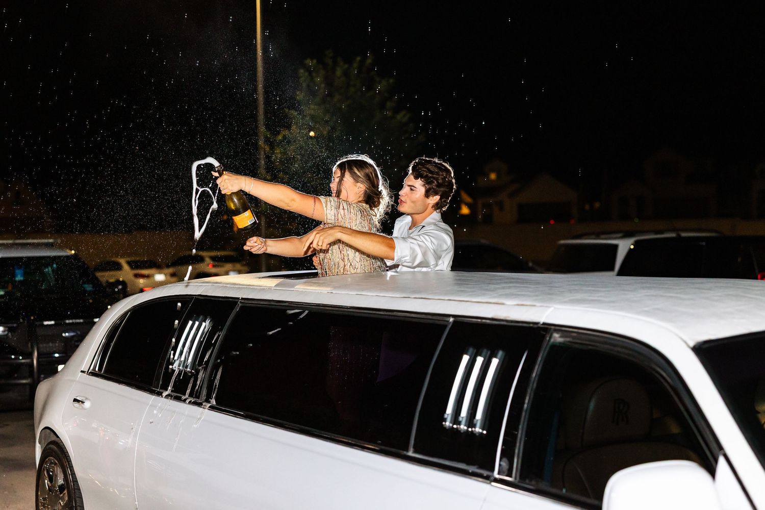 Two people celebrate in a white stretch limousine sunroof, popping champagne at night.