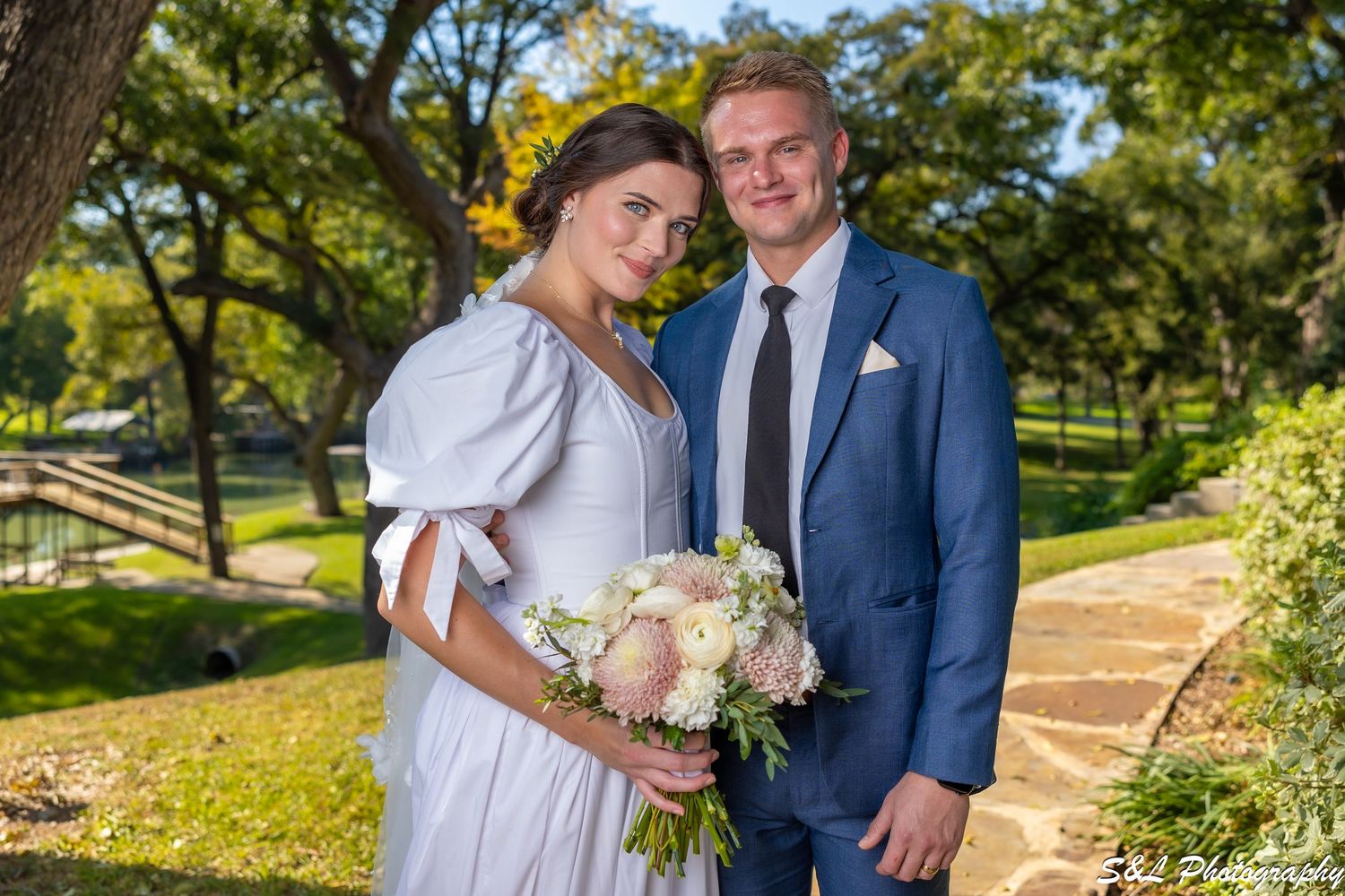 Outdoor wedding portrait in a garden setting with a bouquet of pink and white flowers.