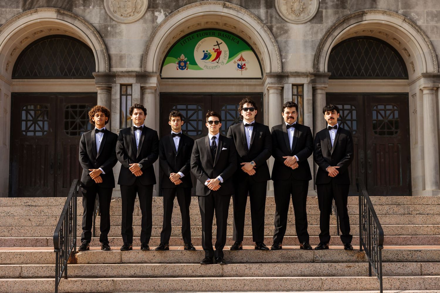 A group of groomsmen in black tuxedos and bow ties stand on stone church steps for a formal wedding photo.