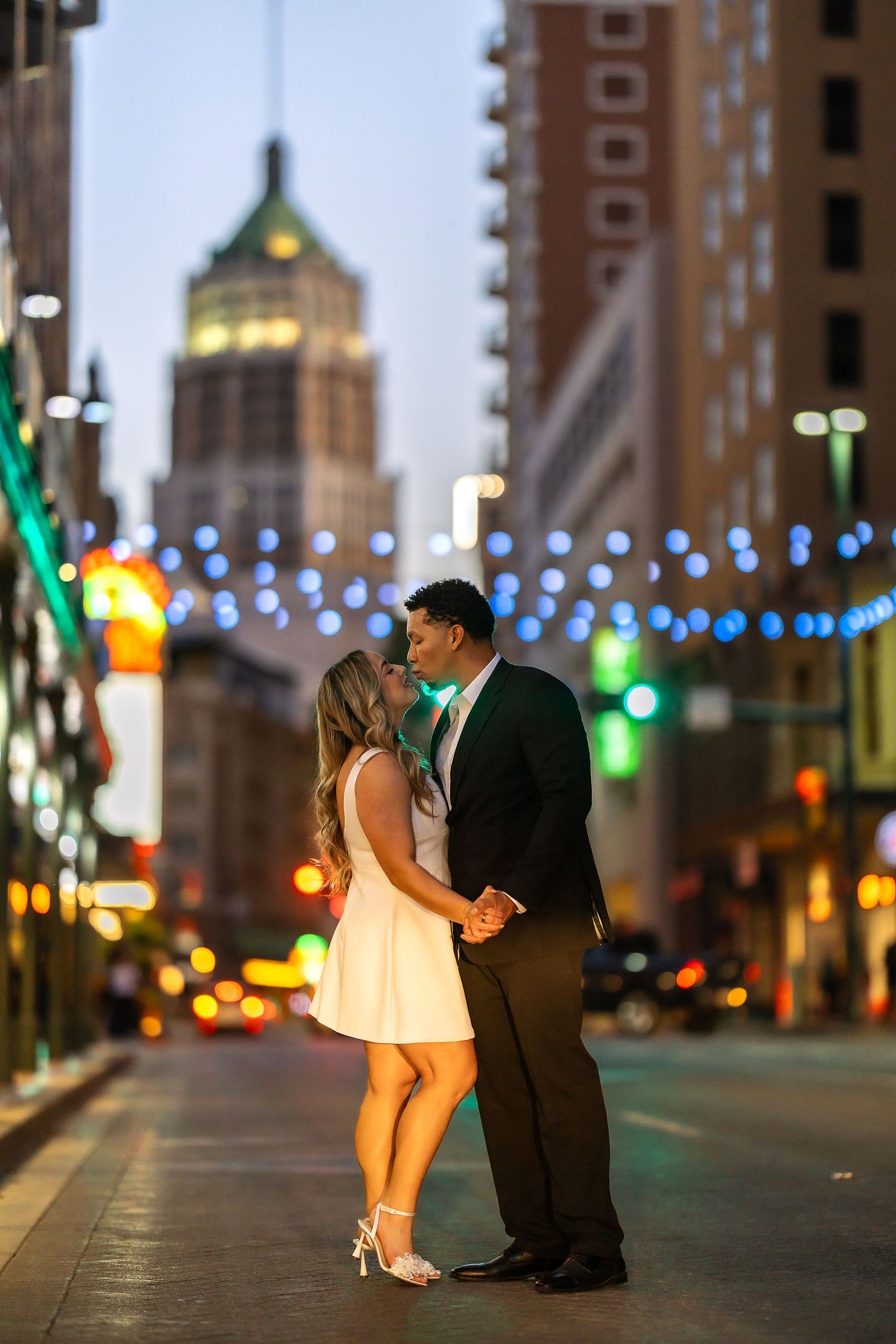 A couple shares an intimate moment on a city street at night with colorful lights in the background.