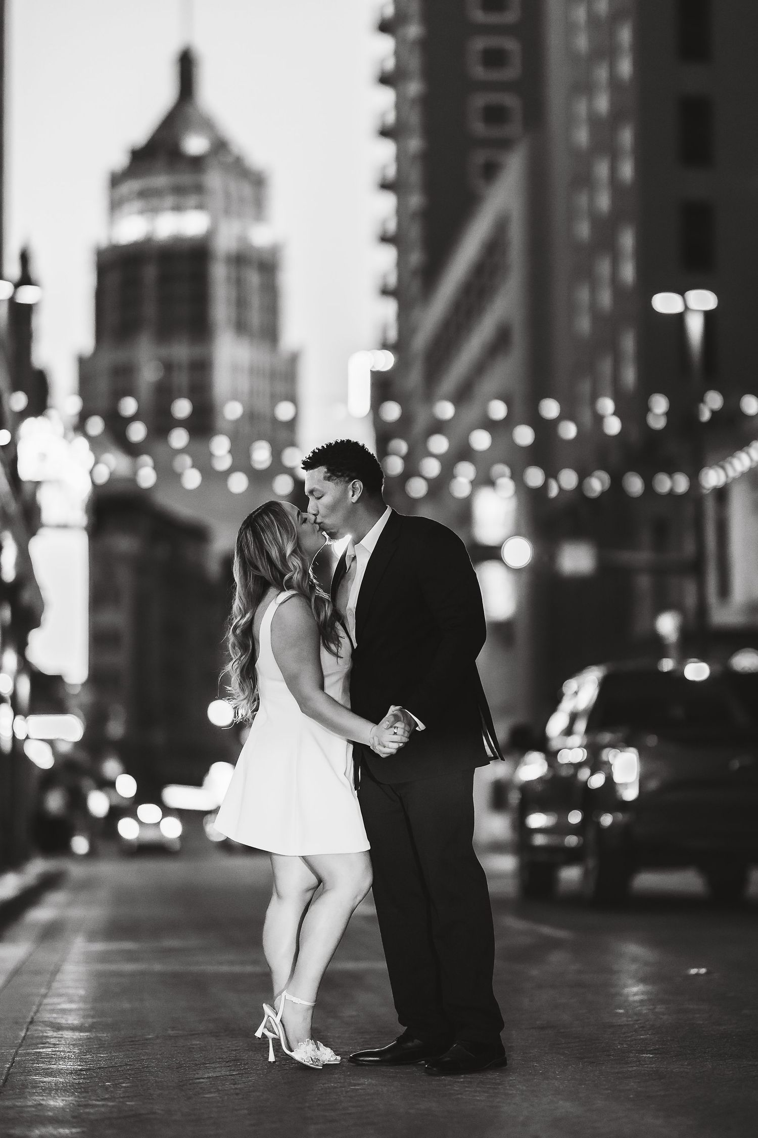 Romantic couple dances together on city street at night with twinkling lights in the background.