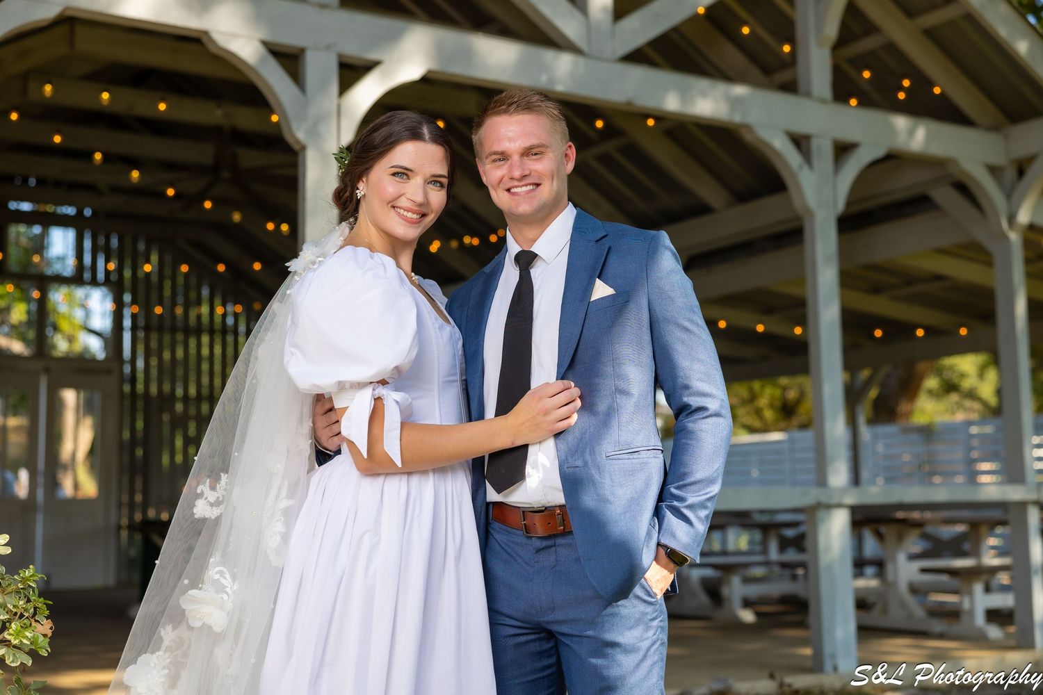 A couple celebrates their wedding day under a rustic wooden pavilion with twinkling lights.