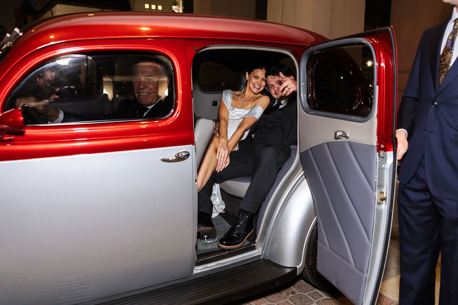 Newlyweds sit inside vintage red and silver car during wedding celebration.