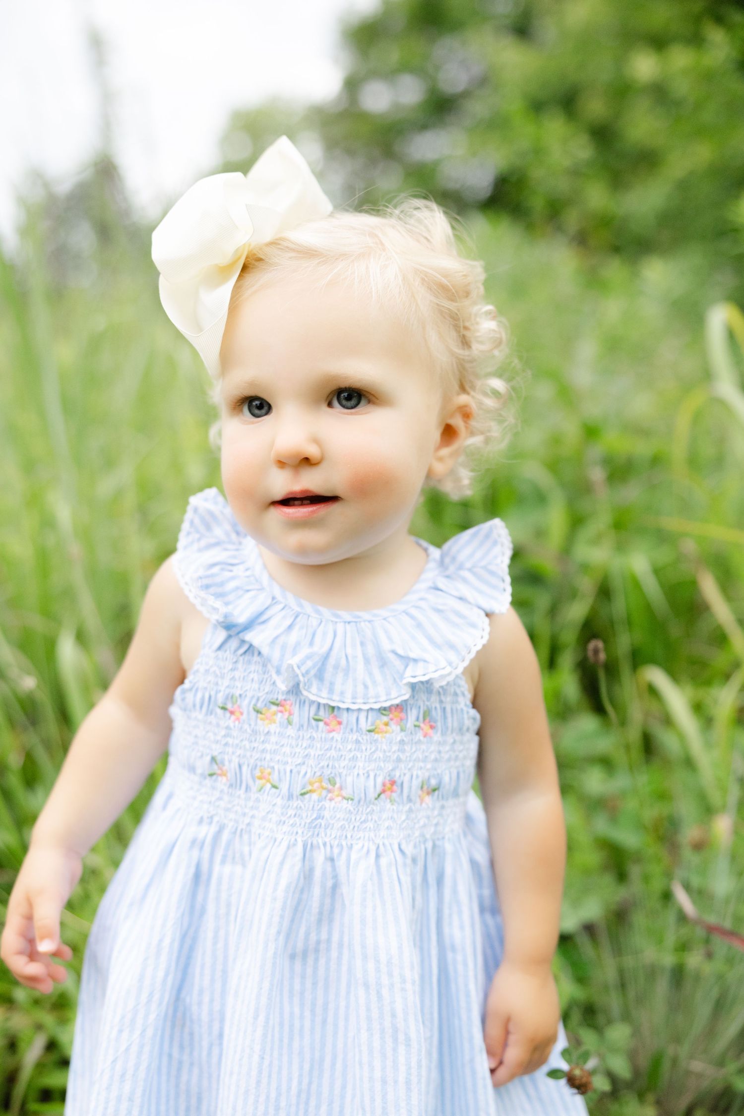 Toddler girl in blue smocked dress with yellow bow headband standing outdoors in green field.